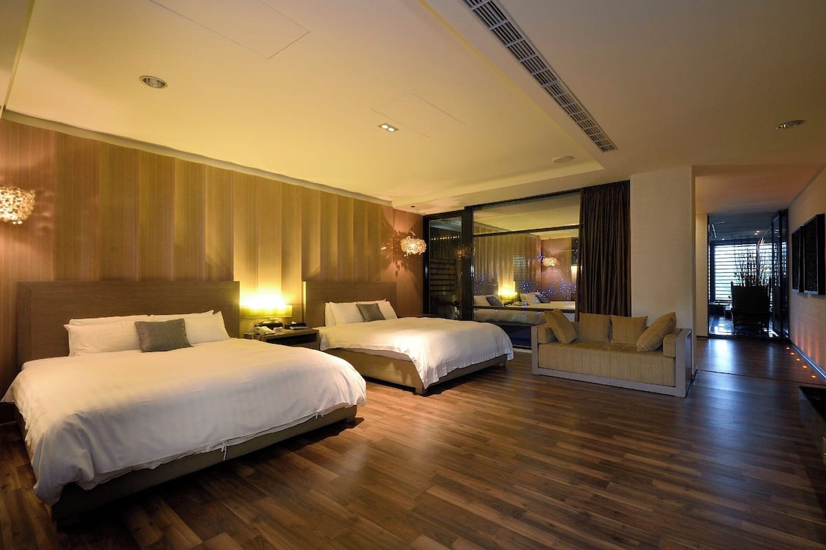 A modern hotel room with two beds, a sofa, and a large window with a city view.