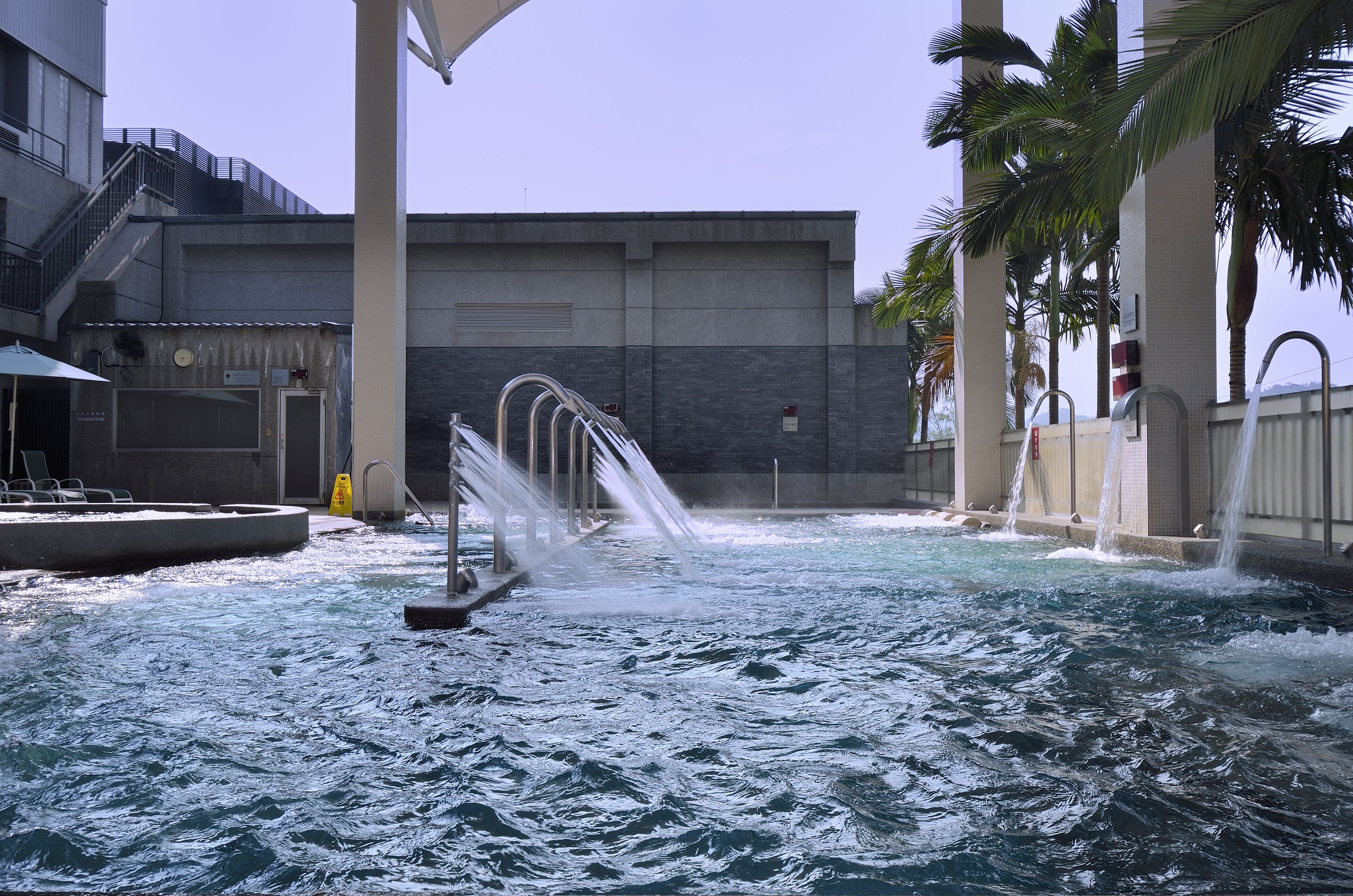 Una fuente ornamental con múltiples fuentes en una zona de piscina, rodeada por un edificio moderno con una escalera y palmeras.