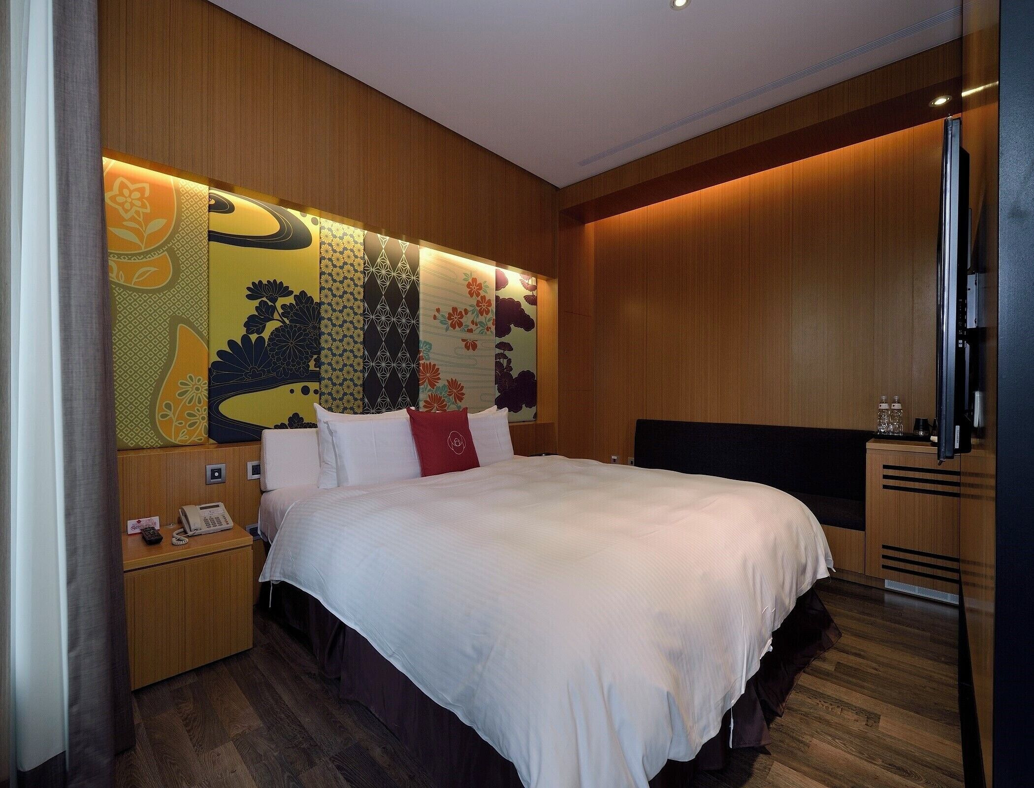 Une chambre d’hôtel avec un grand lit, une tête de lit en bois, un téléviseur à écran plat, une table de chevet avec un téléphone et un mur recouvert d’un papier peint à motifs.