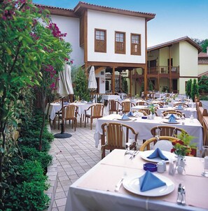 An outdoor dining area with tables set for guests, surrounded by lush greenery and a building with multiple windows. - Tekeli Konakları (Antalya)