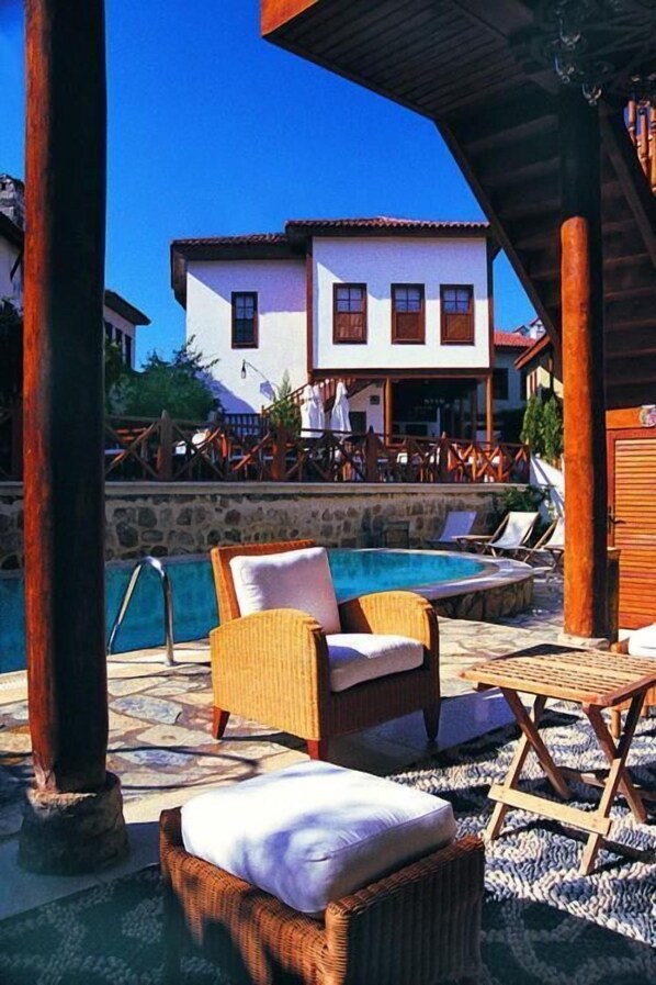 A poolside area with wicker furniture and a stone floor, a pool, and a building with a white facade and red roof in the background.