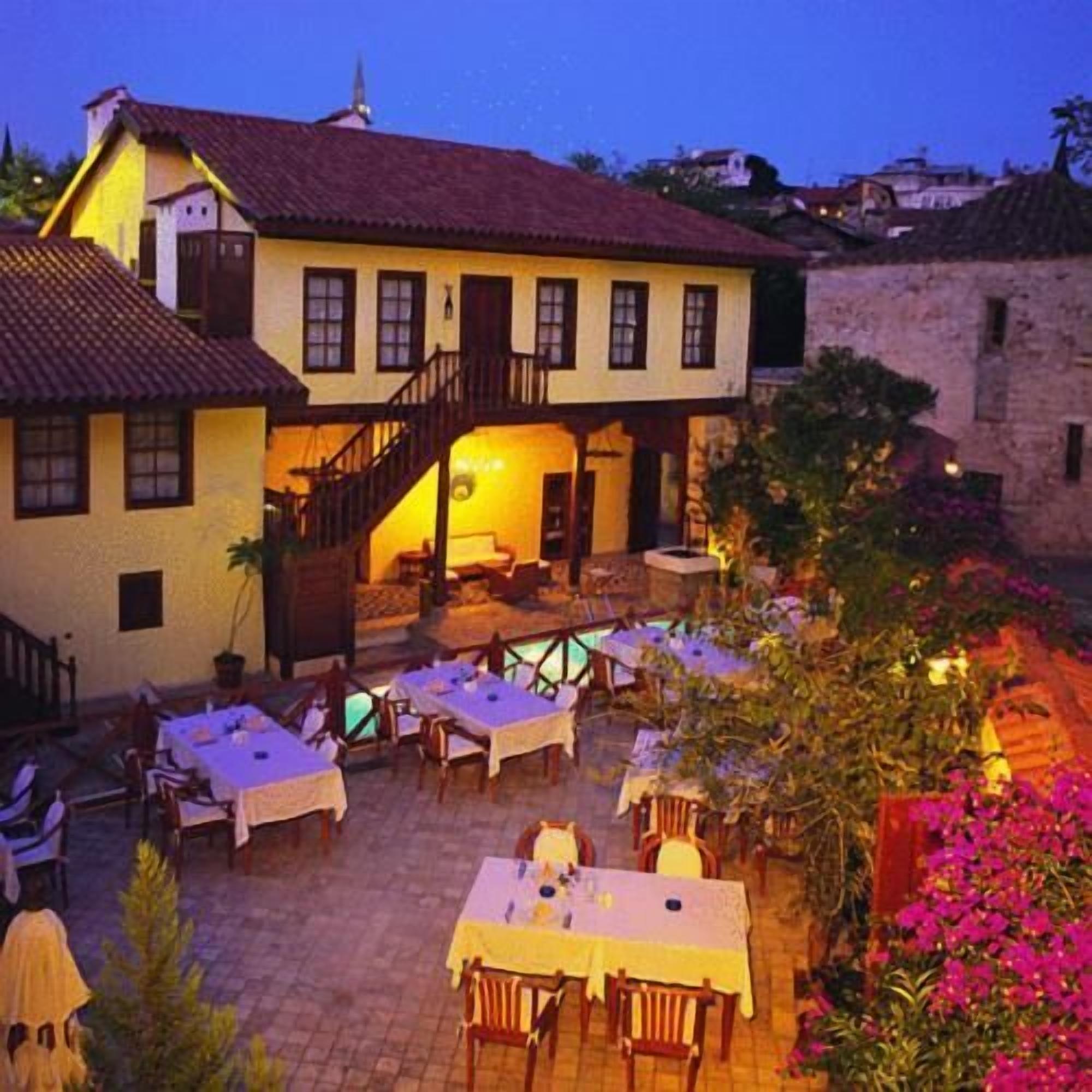 Un edificio con una escalera que lleva a un balcón, zona de comedor al aire libre con mesas y sillas, y un jardín con flores.