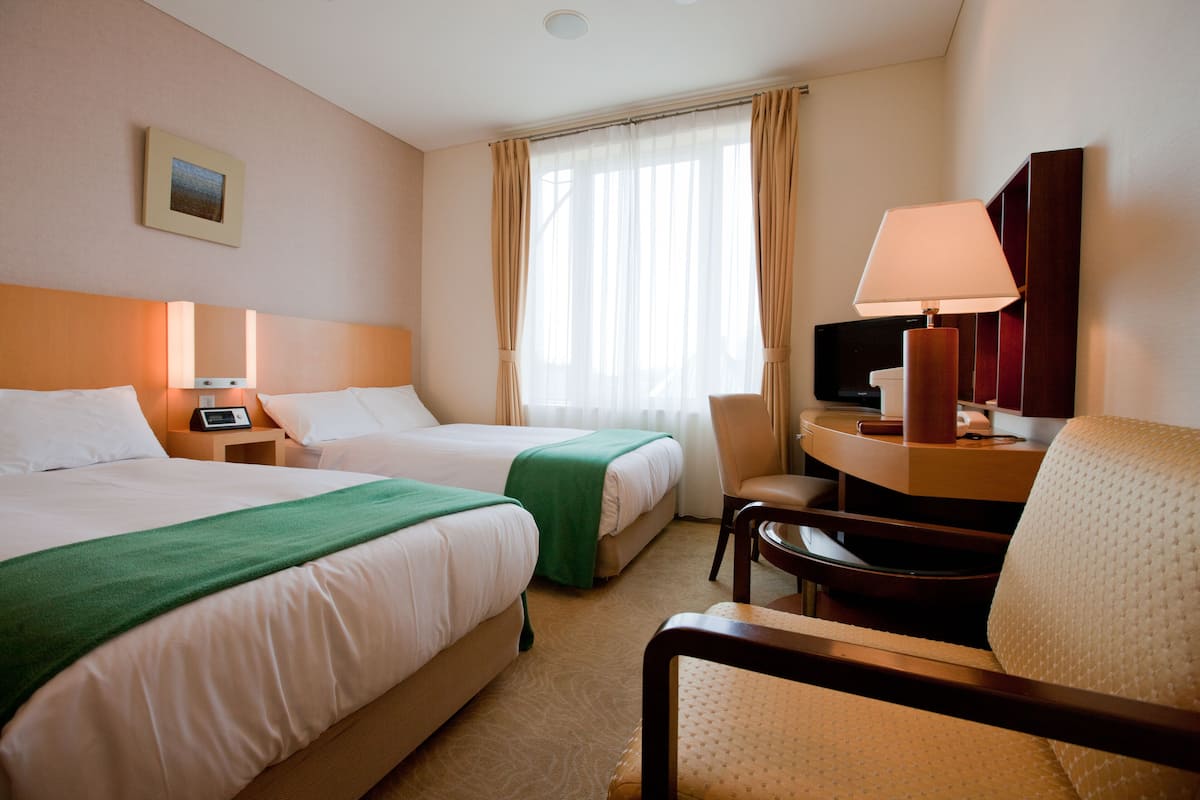A hotel room with two beds, a desk with a lamp, a chair, a TV, and a painting on the wall.