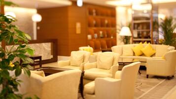 A modern hotel lobby with a seating area featuring cream-colored sofas and armchairs, a coffee table, and a bookshelf with books.