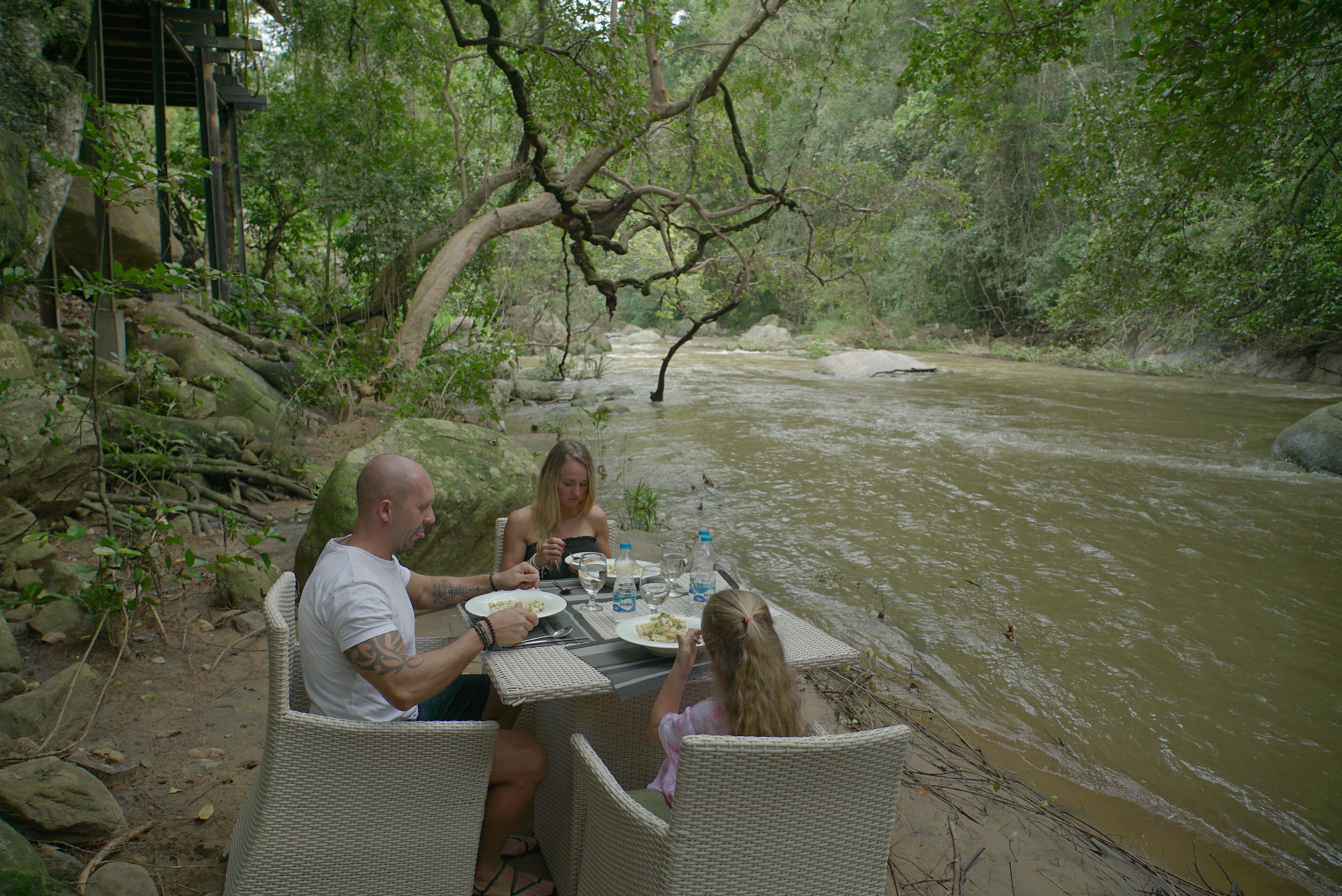 A dining table set up by a river with wicker chairs, surrounded by greenery and rocks.