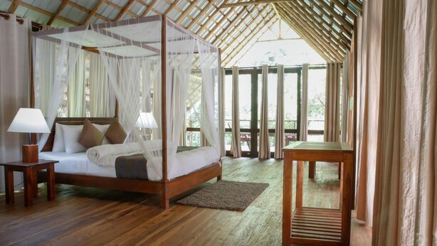 A four-poster bed with white linens and a canopy, a wooden nightstand with a lamp, and a small table with a lamp.