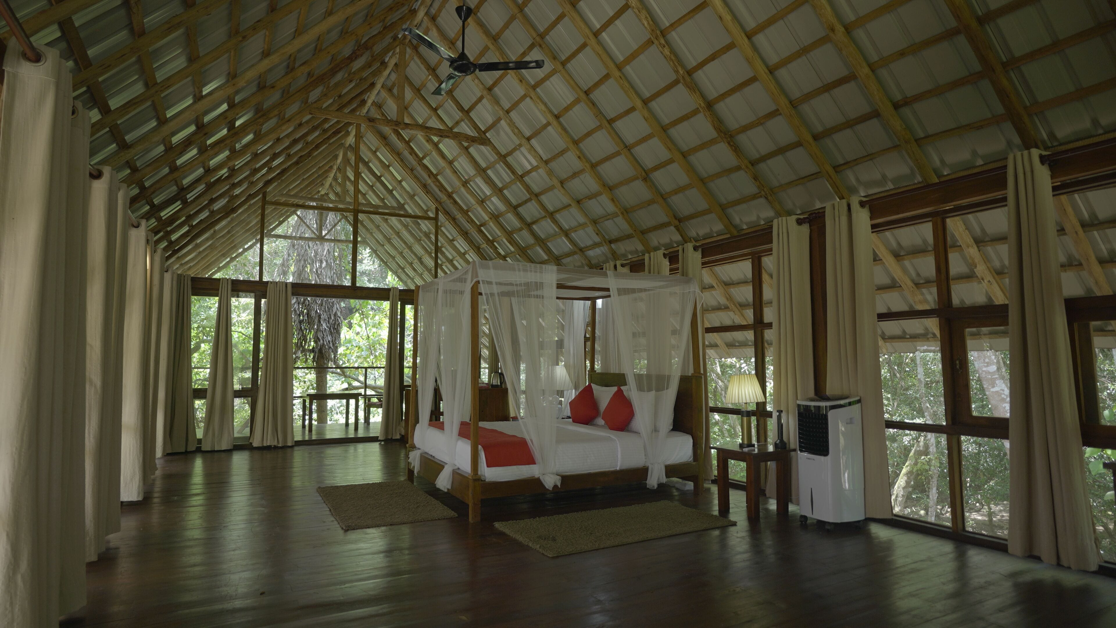 A spacious room with a wooden floor, a four-poster bed, and large windows offering a view of the outdoors.