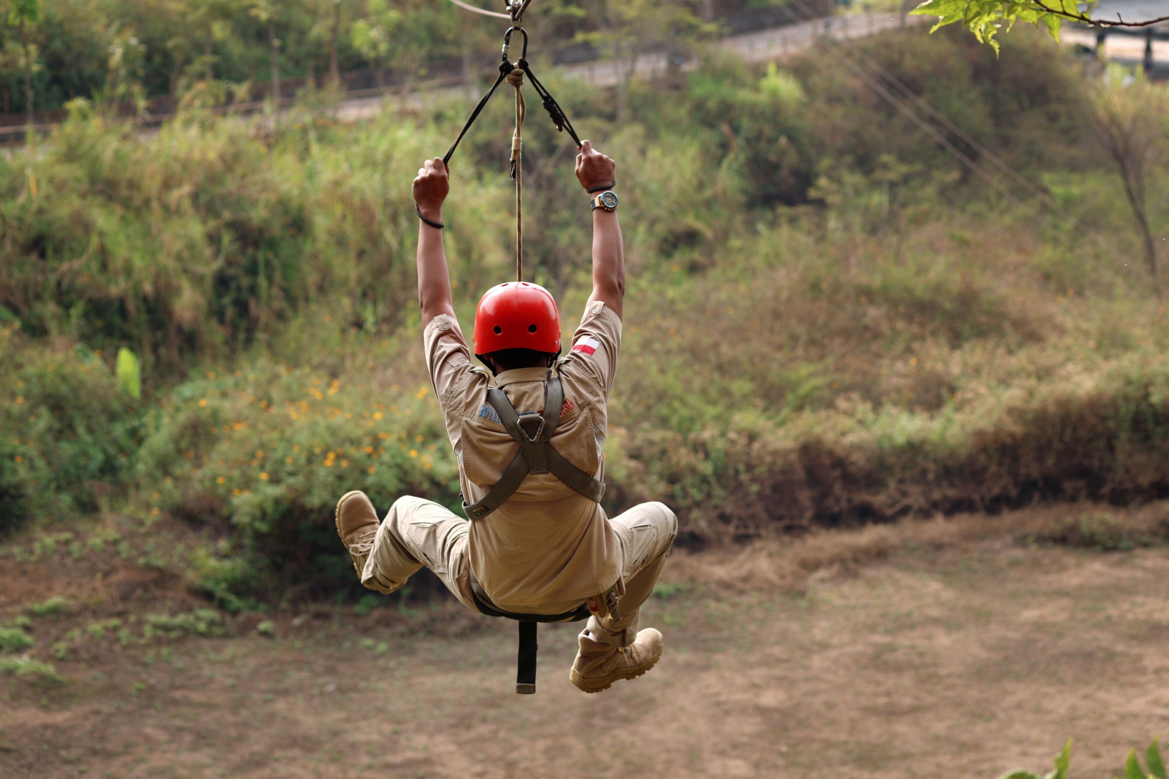 A person suspended in mid-air, wearing a red helmet and harness, with a natural landscape in the background.