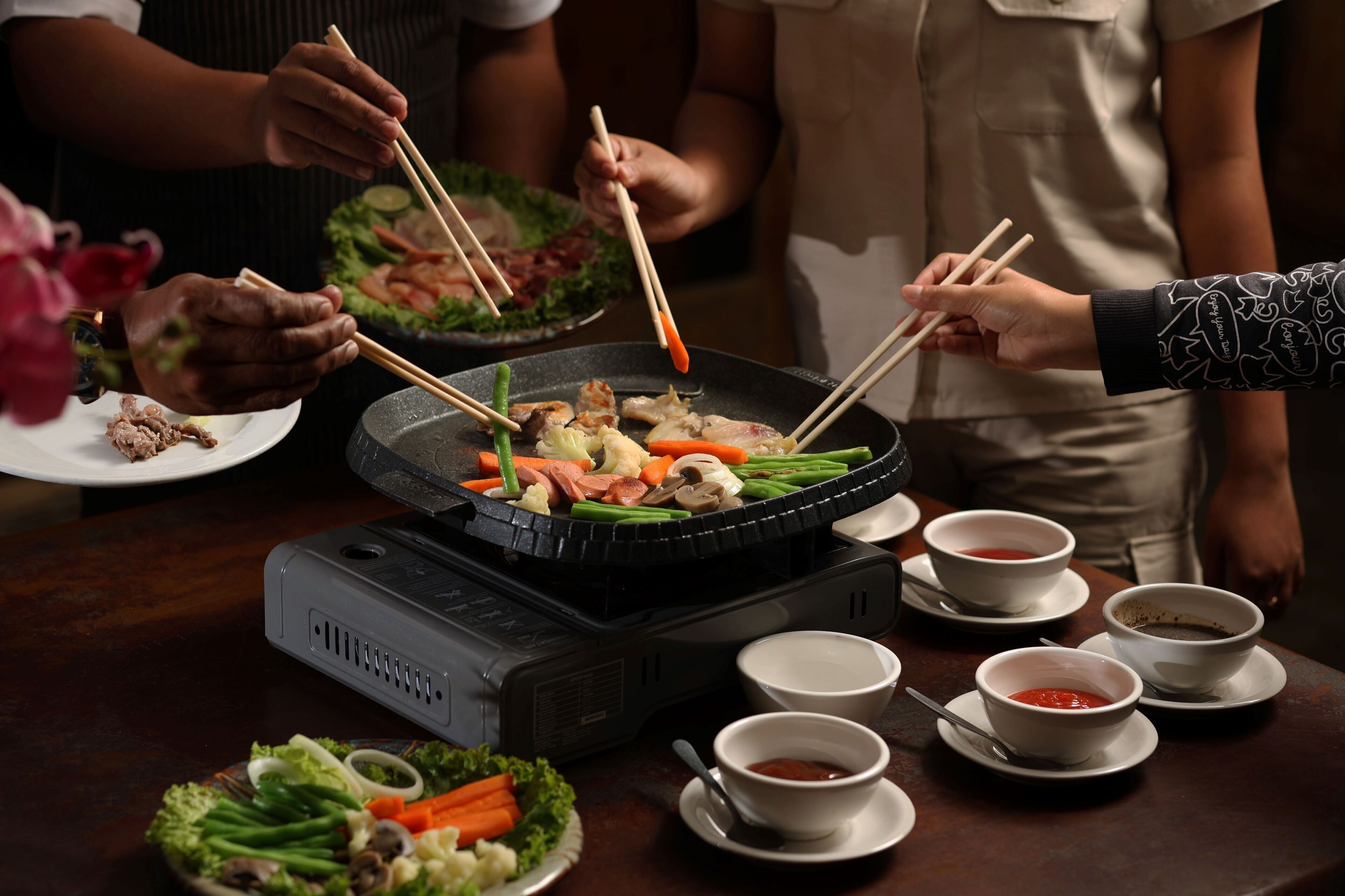 A hot pot setup with various vegetables and meats, surrounded by bowls of sauces and a portable heater.