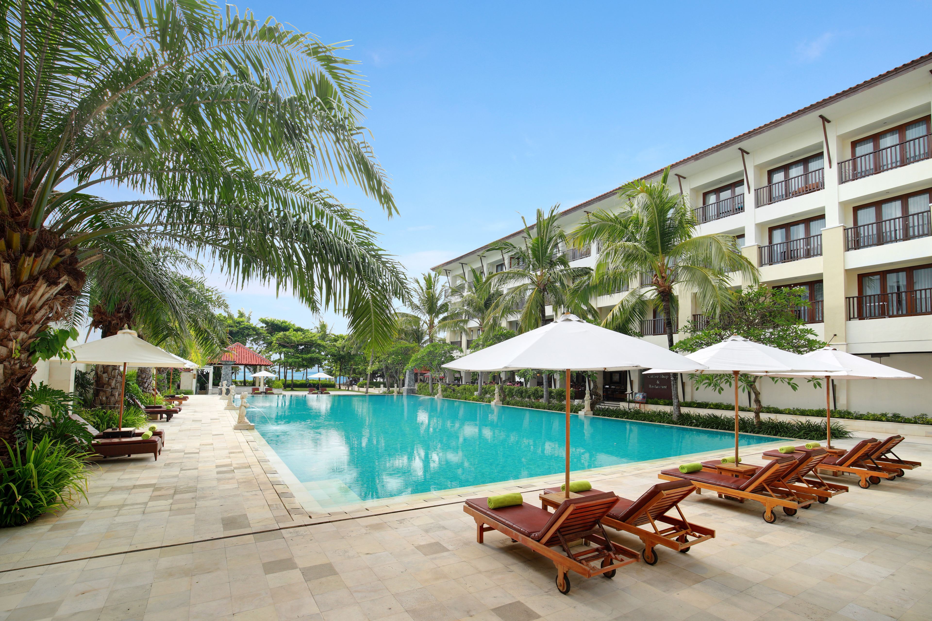 A pool area with lounge chairs and umbrellas, surrounded by palm trees and a building with balconies.