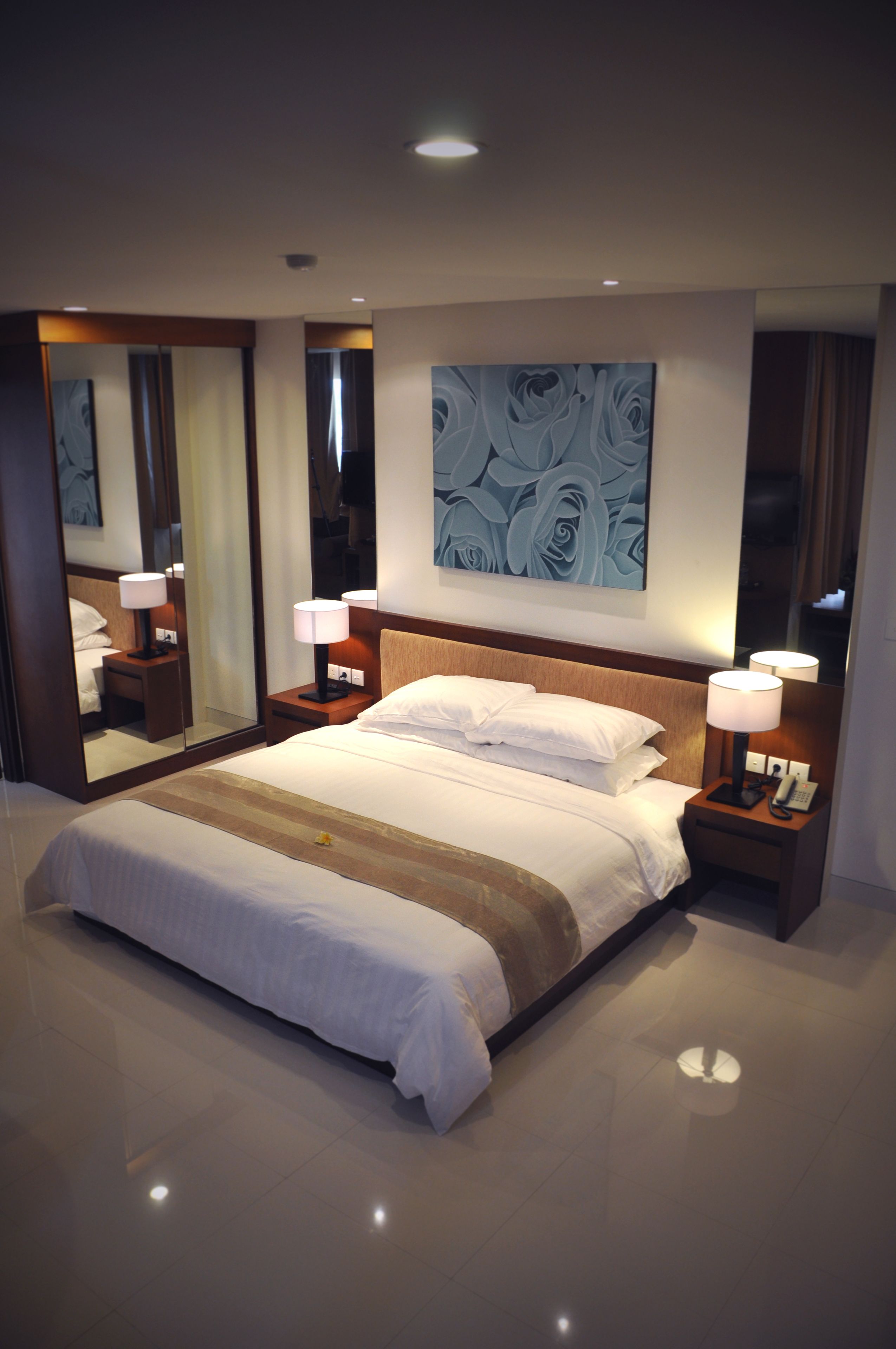 Ein modernes Hotelzimmer mit einem großen Bett, zwei Nachttischen inklusive Lampen, einem großen Spiegel und einem an der Wand befestigten abstrakten Kunstwerk.