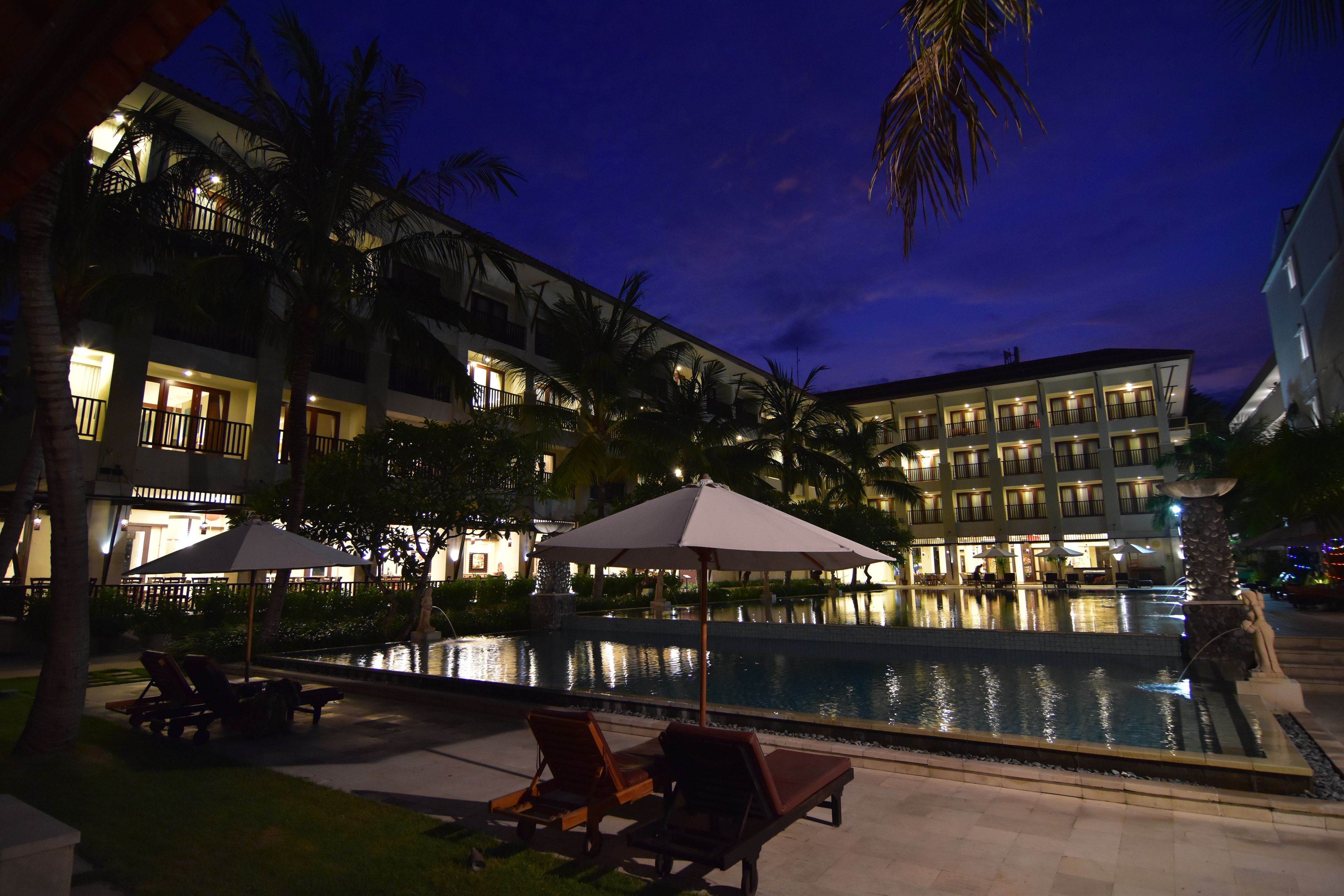 Vista nocturna de una piscina de hotel con sillones de descanso y sombrillas, rodeada de palmeras y edificios iluminados.