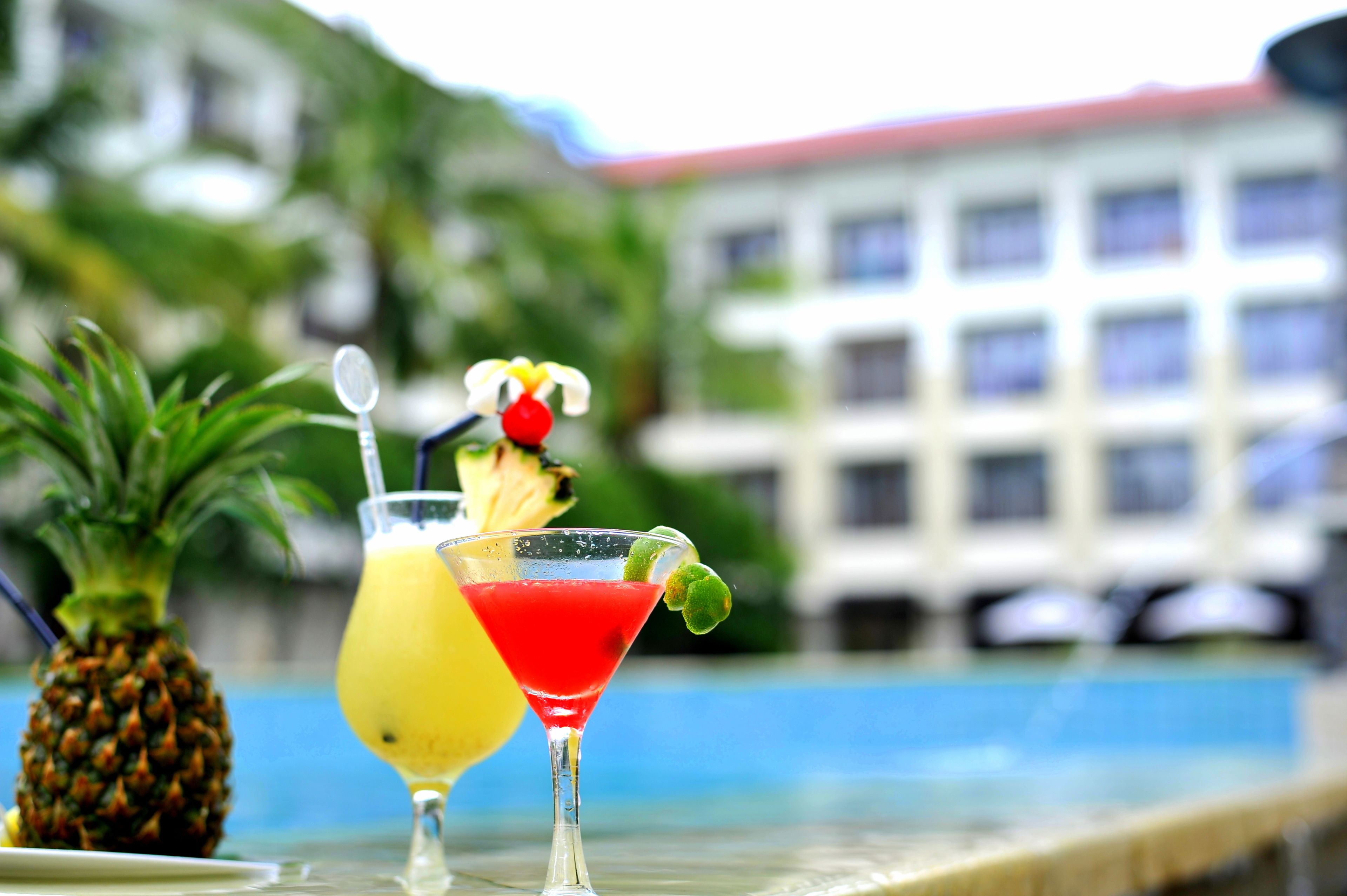 Two cocktails with a pineapple and a lime on the rim, placed on a table by a poolside with a building in the background.