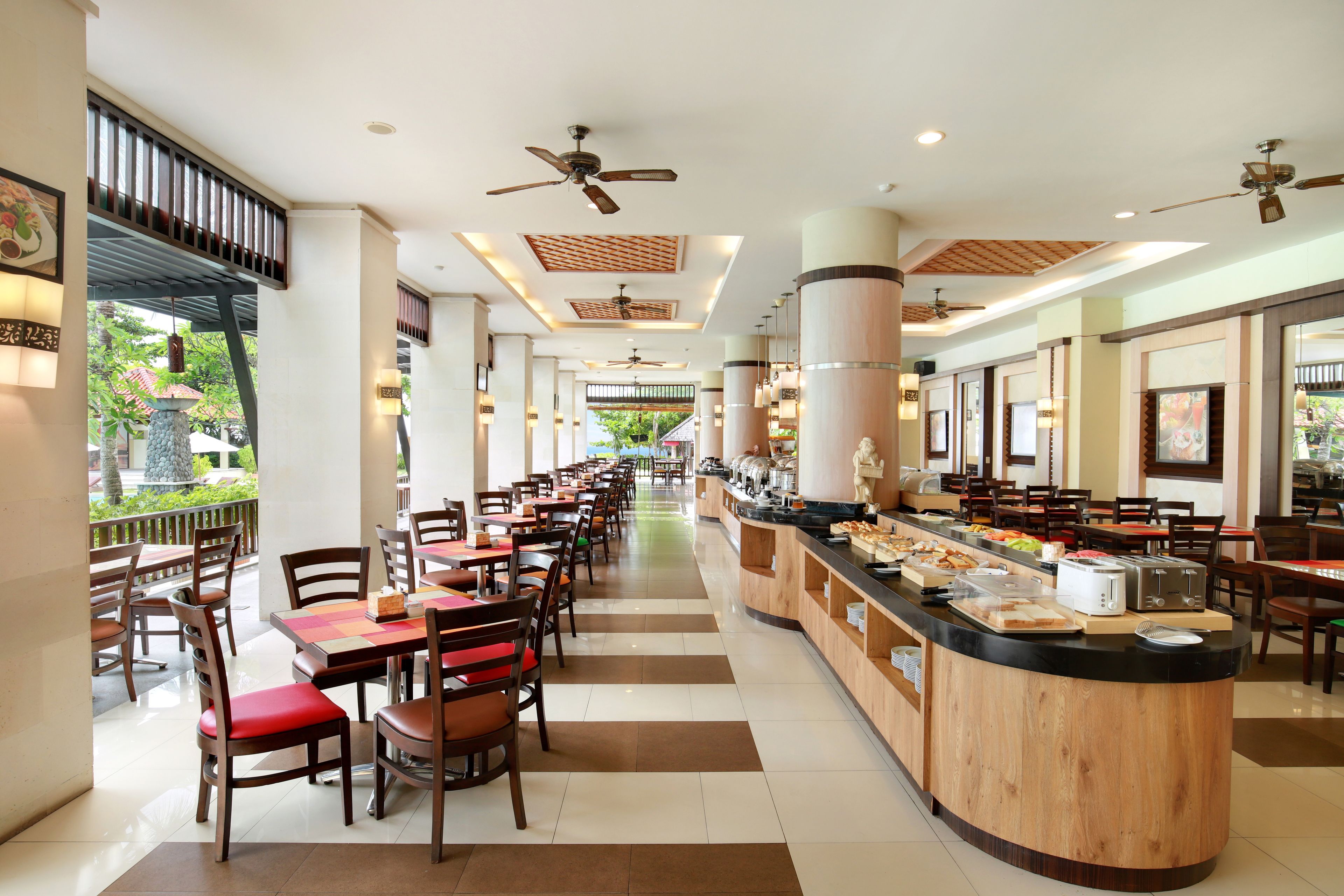 A spacious hotel dining area with multiple tables and chairs, a central buffet counter, and ceiling fans.