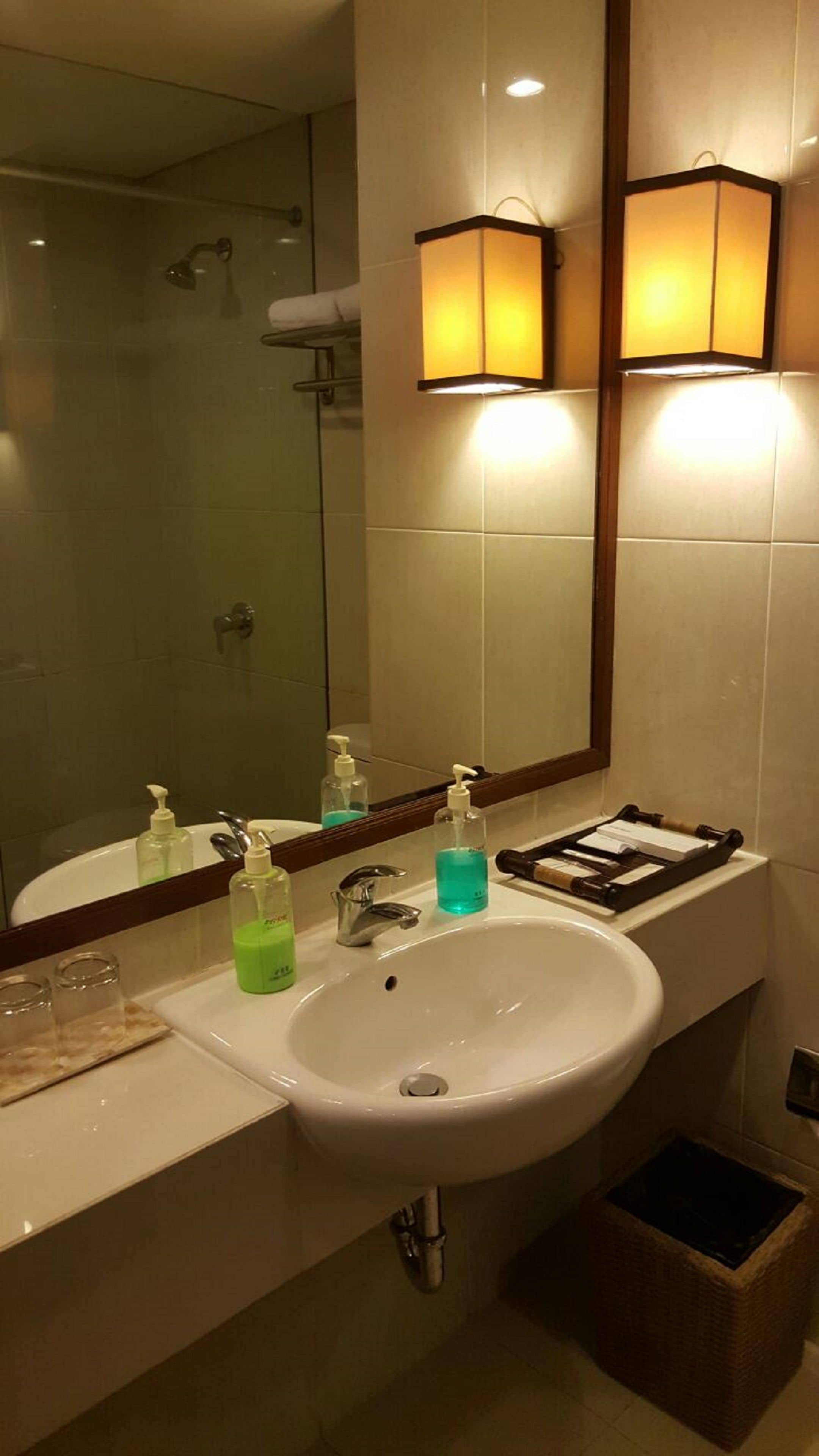 Un baño de hotel con un lavamanos blanco, dos lámparas fijadas a la pared, un espejo y una zona de ducha.