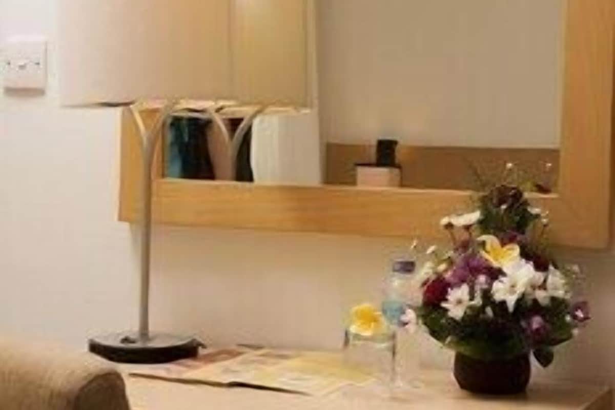 A small desk with a lamp, a mirror, a vase with flowers, and a bottle.