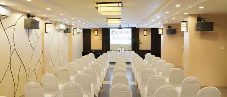 A conference room with rows of white chairs, a long table in the center, and a projection screen at the front.