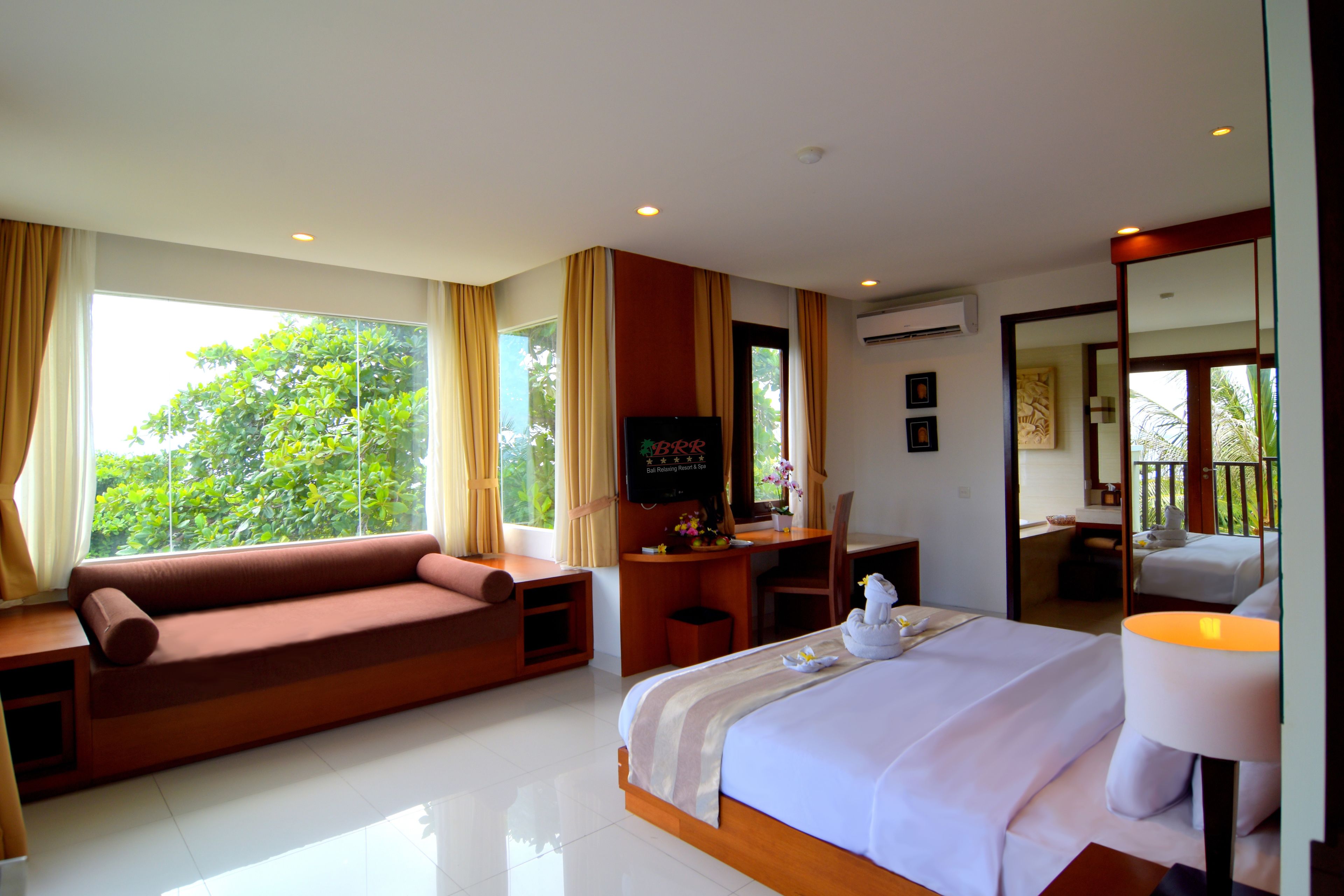 A hotel room with a bed, a desk, a sofa, and a view of greenery outside.