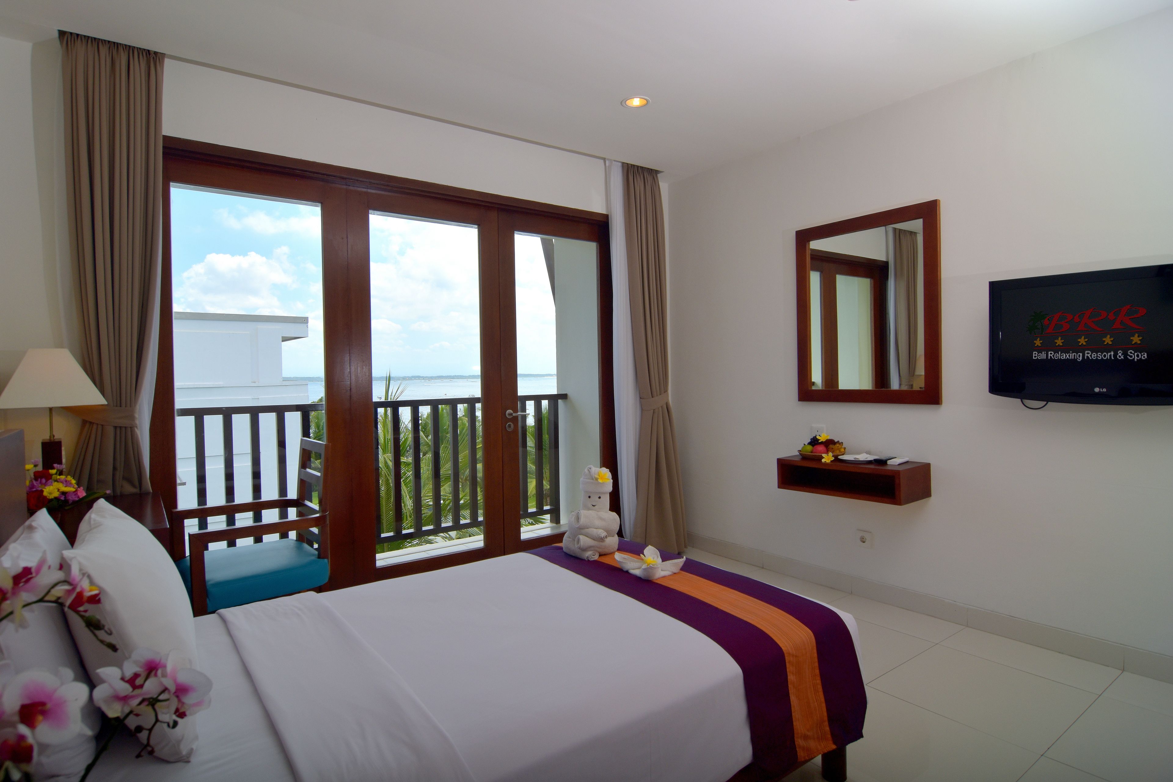 Habitación de hotel con cama, televisión, balcón y vista al mar, además de una silla de madera.