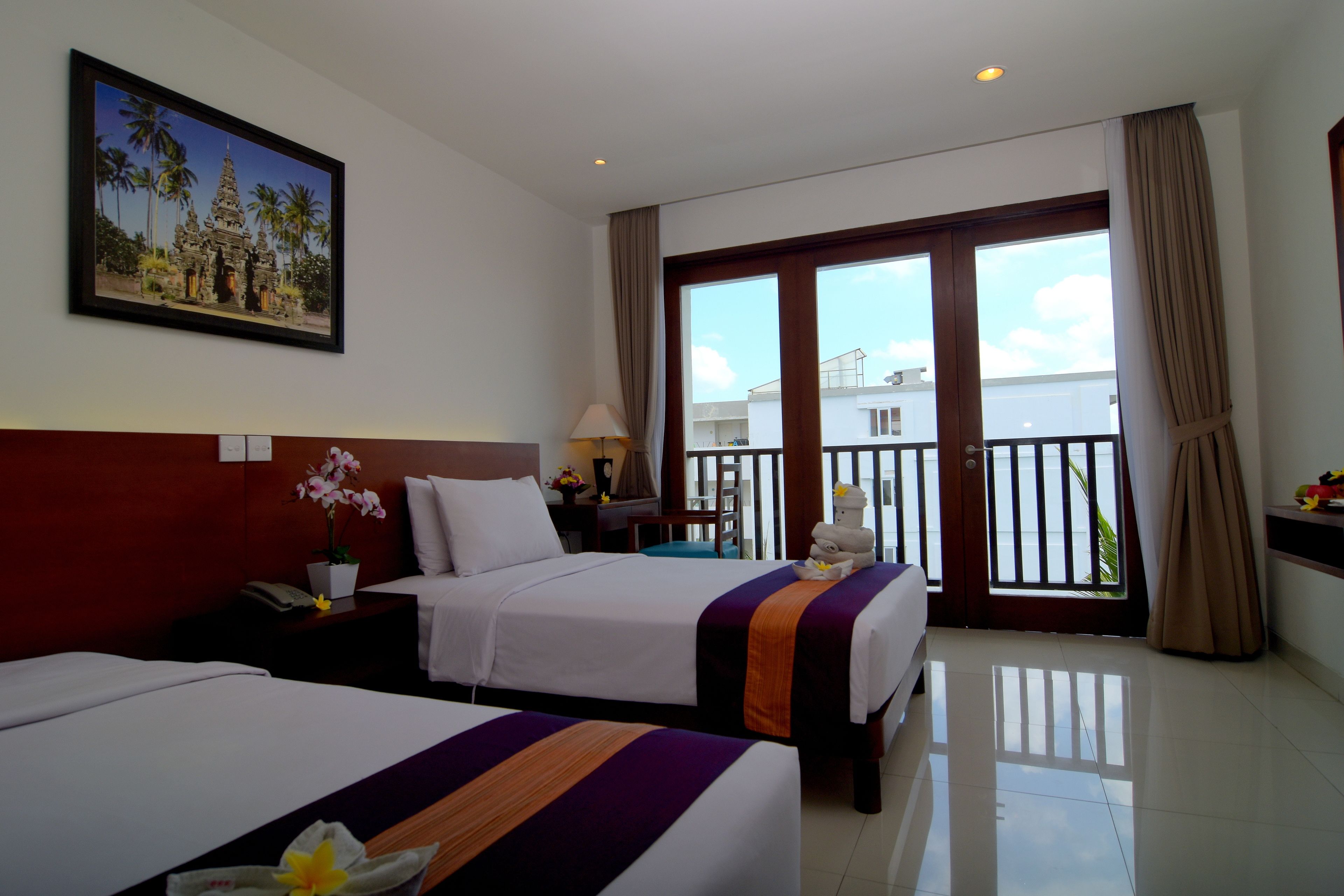 A hotel room with two beds, a balcony with a view, a painting on the wall, and a desk with a lamp.
