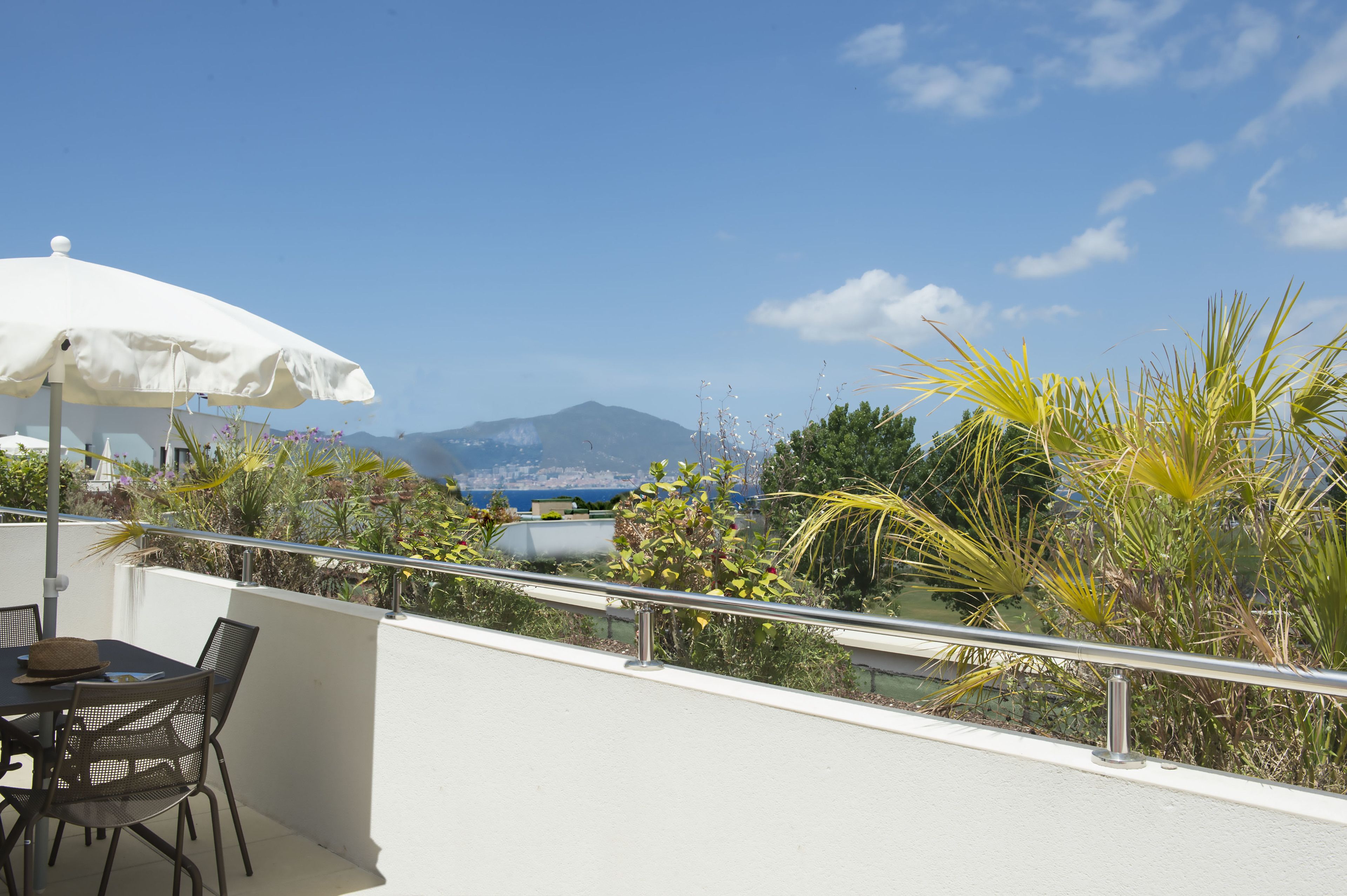 Un balcon avec une table et des chaises, un parasol blanc, et une vue sur la mer et les montagnes.