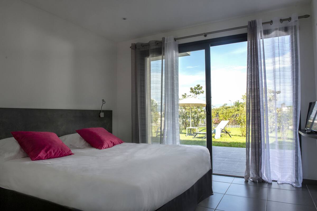 Ein Schlafzimmer mit einem großen Bett, zwei rosafarbenen Kissen, einem dunklen Kopfteil und einer Glastür, die auf einen Balkon mit Gartenblick führt.