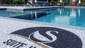 Zona de piscina amb gandules i ombrelles, un edifici amb la inscripció “Suite Home” al fons, i un logotip al costat de la piscina.