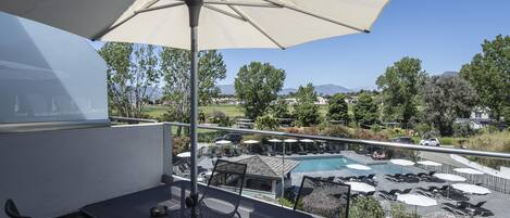 A balcony with a table and chairs, an umbrella, and a view of a swimming pool and surrounding landscape.