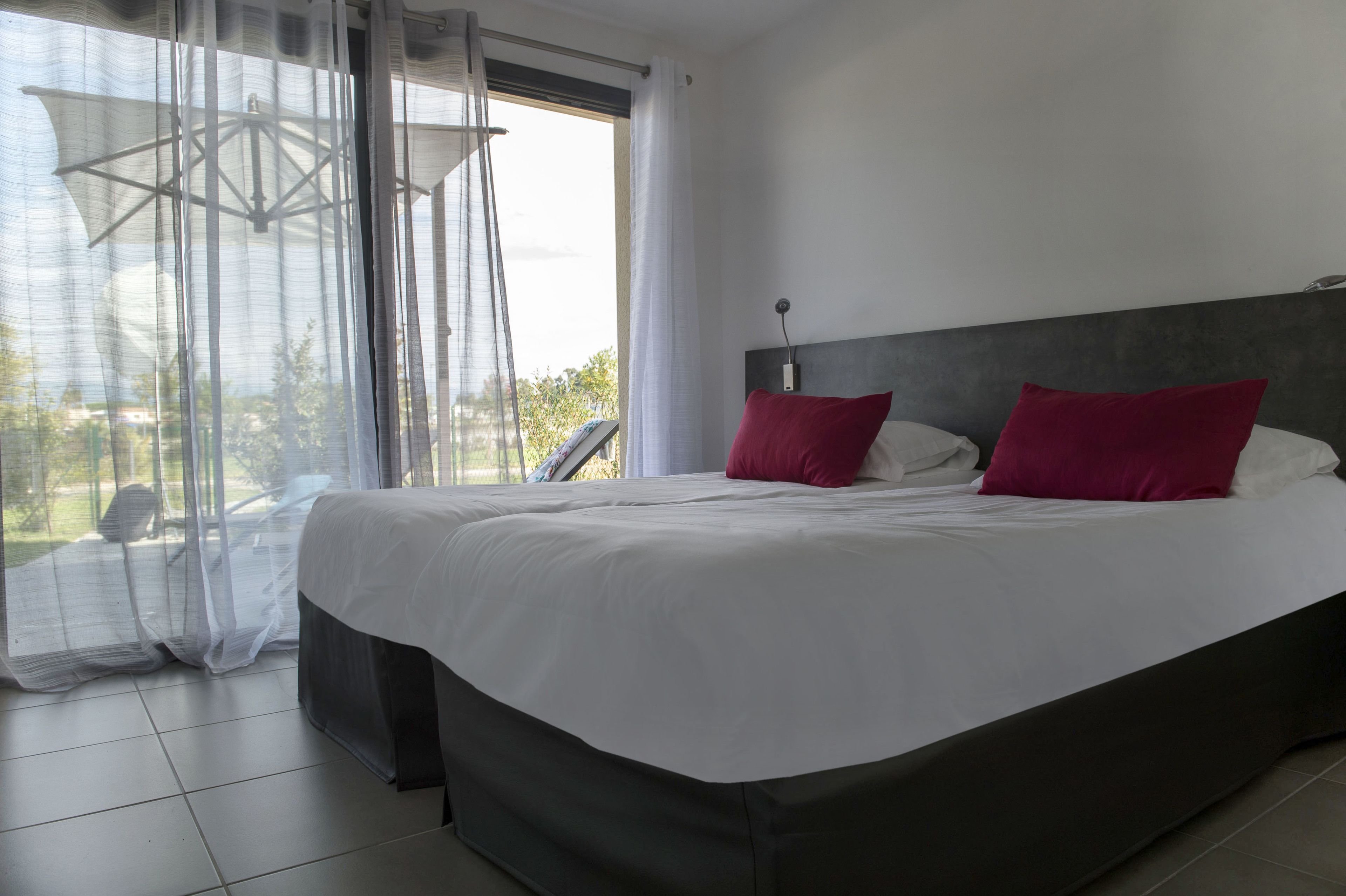 A bedroom with a large bed, a headboard, and a view through sheer curtains.