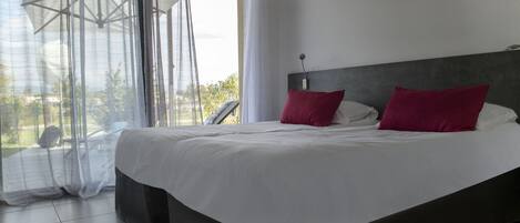 A bedroom with a large bed, a headboard, and a view through sheer curtains.