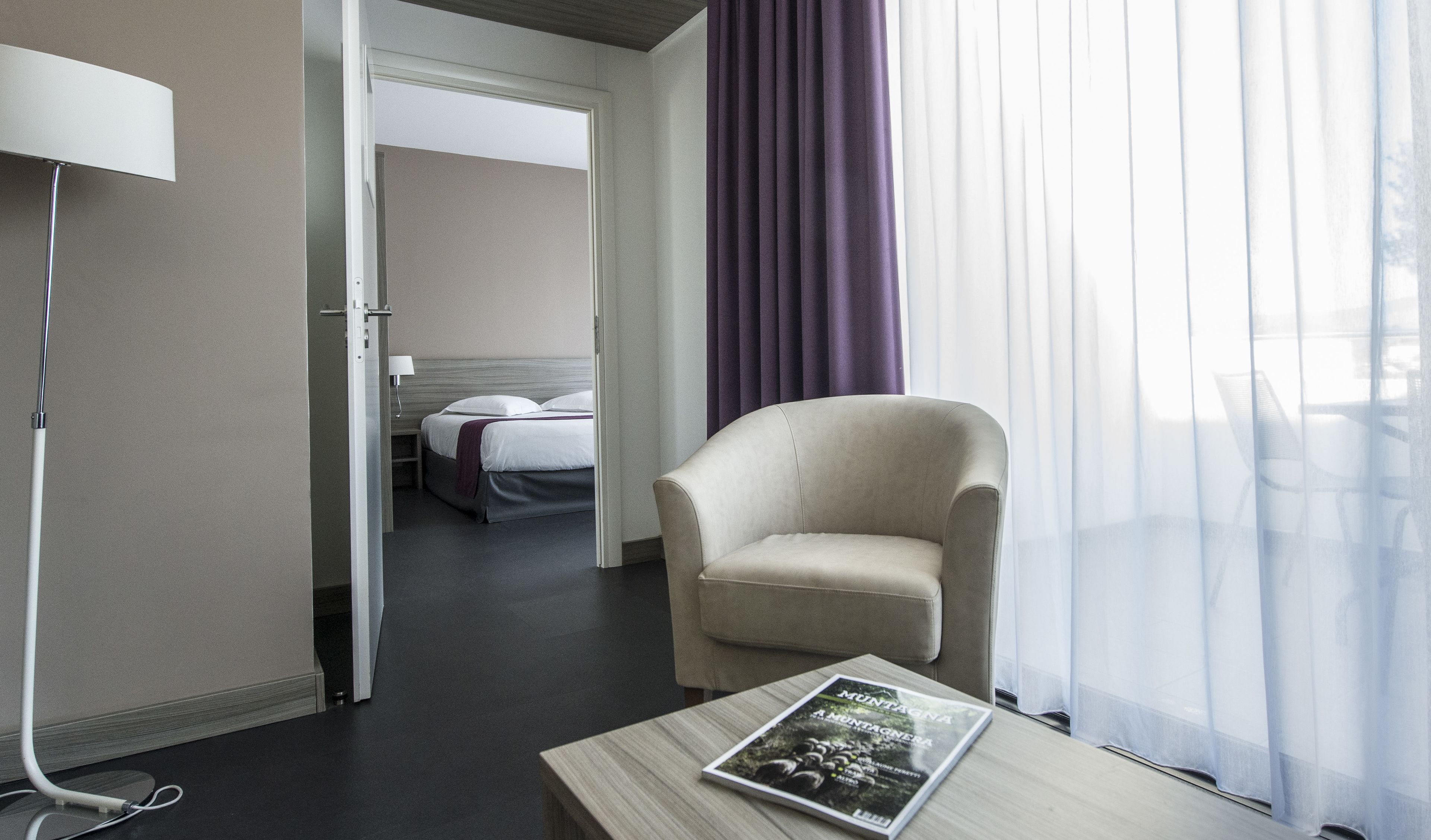 Una habitación de hotel con cama, una silla, una mesa con una revista y una lámpara.