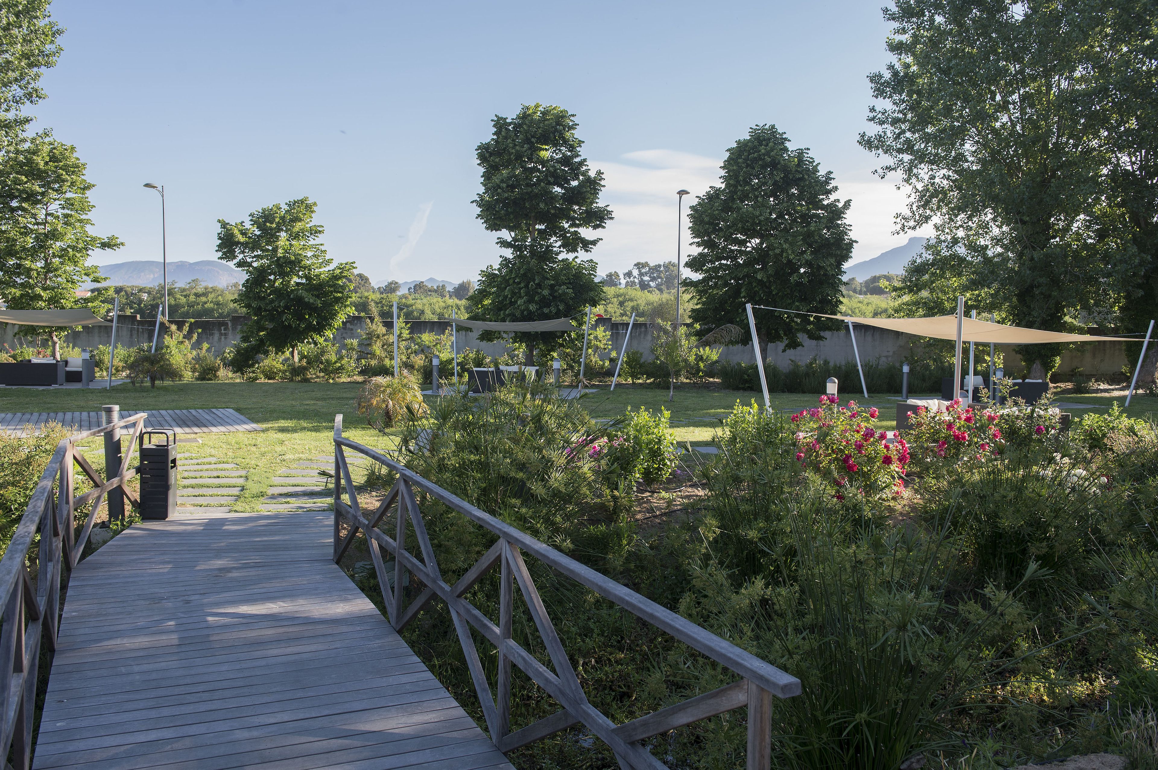 Un pont en bois mène à un jardin doté d’un espace pique-nique, le tout étant entouré d’arbres et avec une montagne en toile de fond.