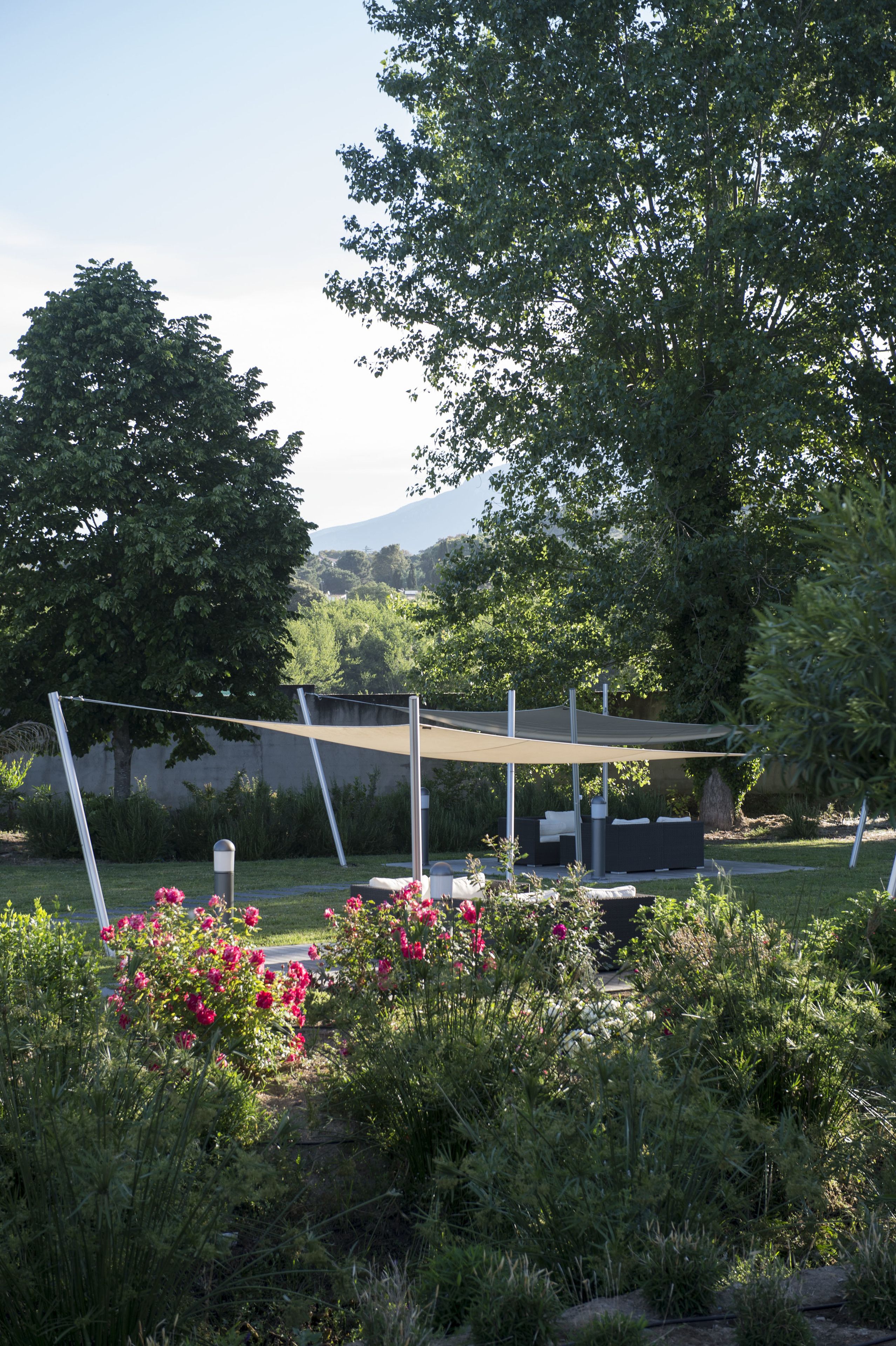 Ein Garten mit Sitzgelegenheit unter einer Sonnenschutzstruktur, umgeben von üppigem Grün und blühenden Pflanzen.