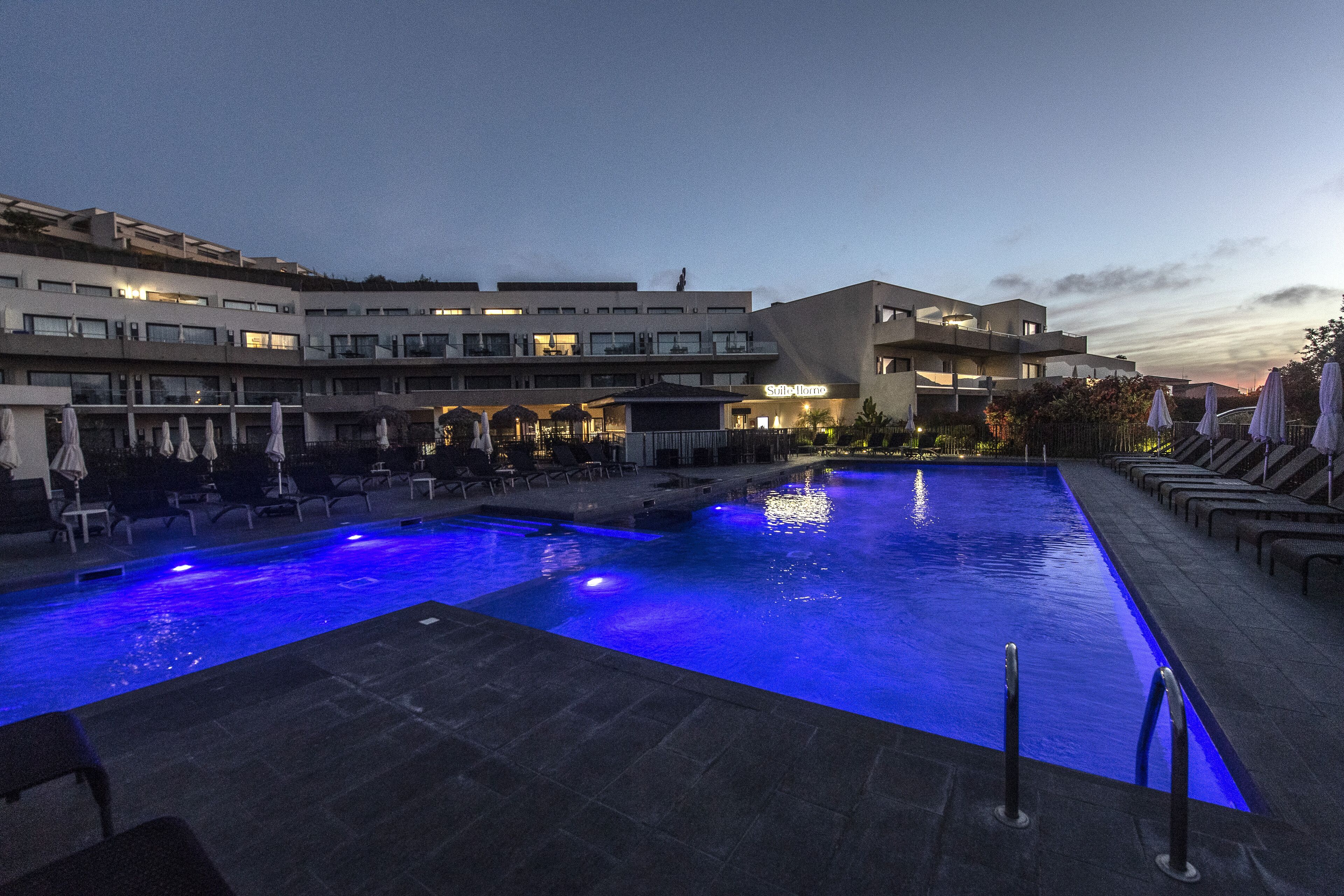 A swimming pool with blue lighting, lounge chairs, and a modern building with multiple floors and balconies.