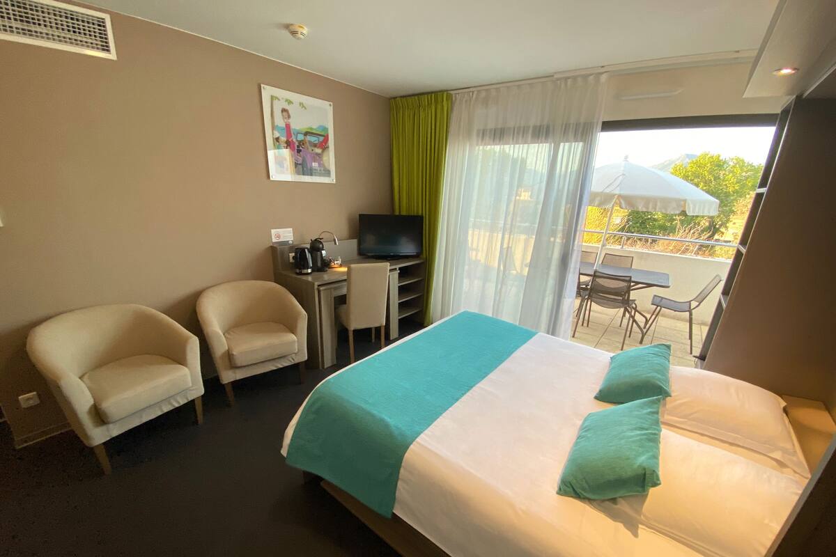 Ein Hotelzimmer mit einem Bett, zwei Sesseln, einem Schreibtisch mit Fernseher und einem Balkon mit Sitzgelegenheit.