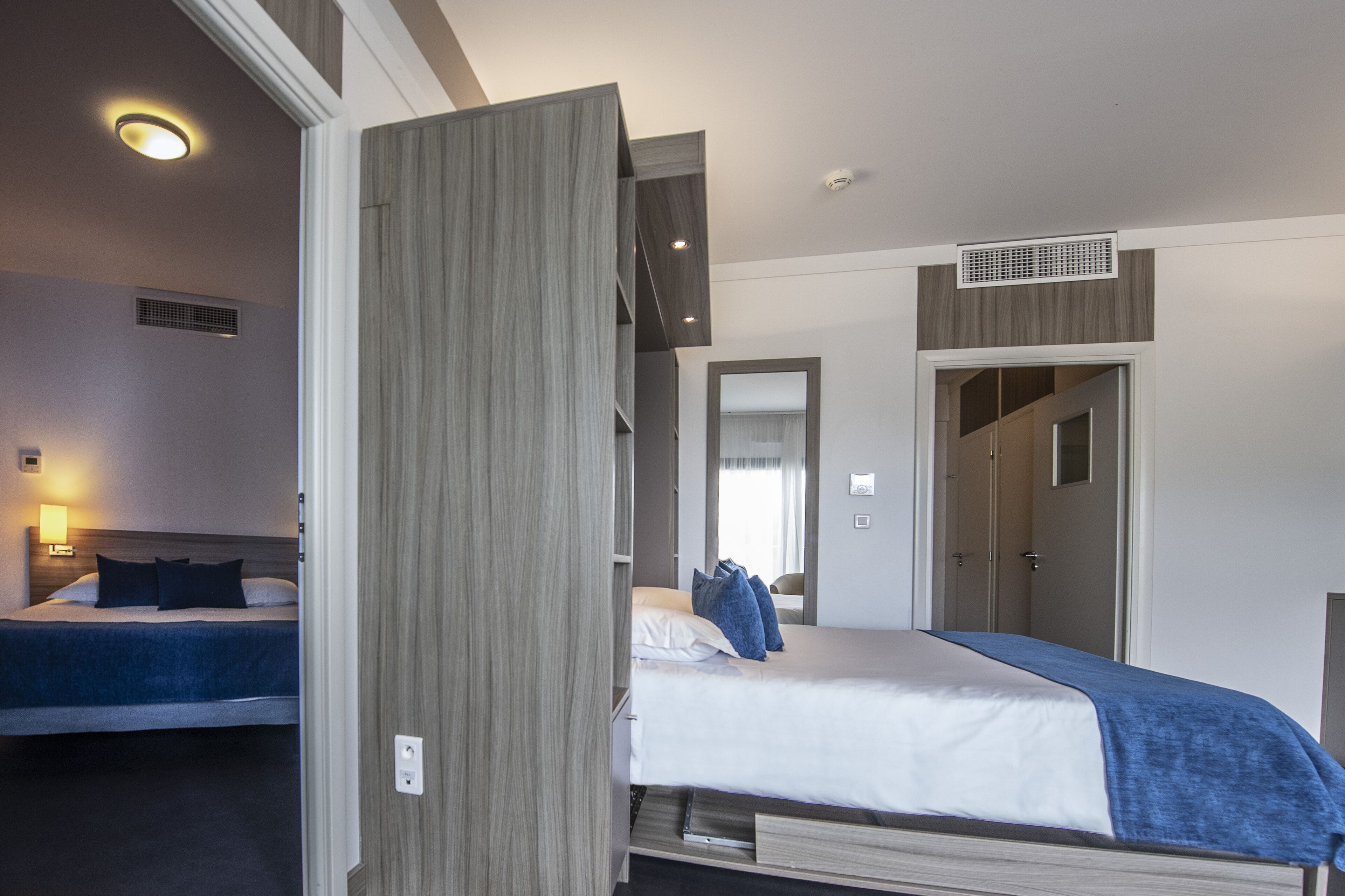 A hotel room with a bed, a wardrobe, a mirror, and a door leading to another room.