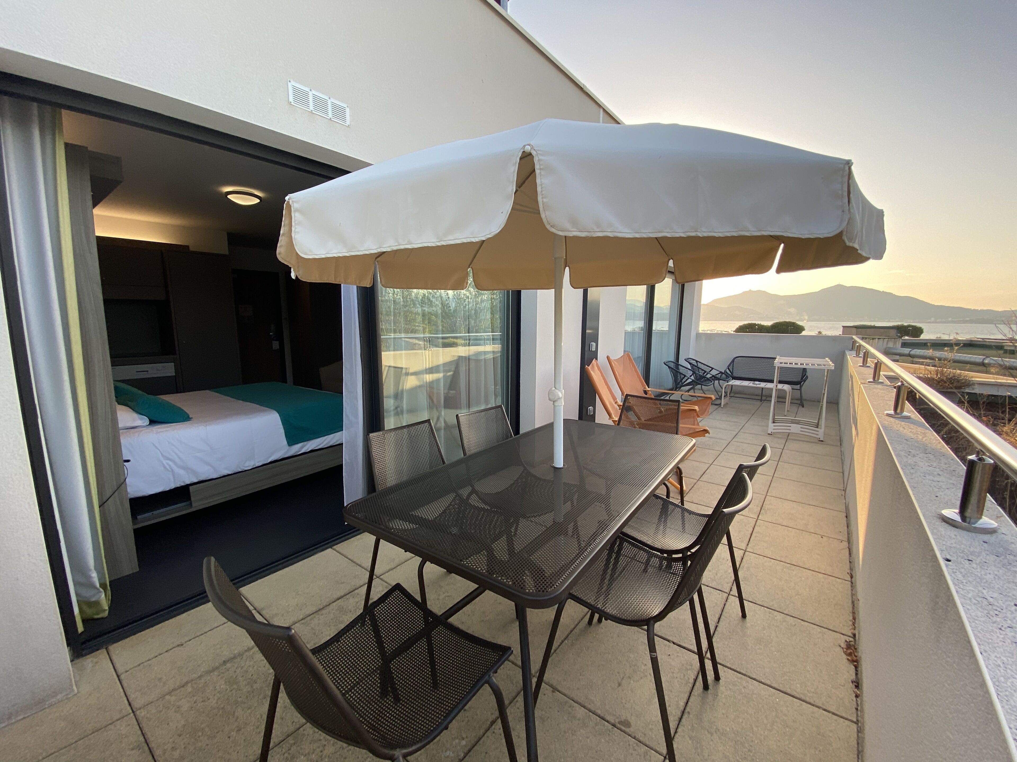 A balcony with a table and chairs, a bed with a green blanket, and a view of the sea.