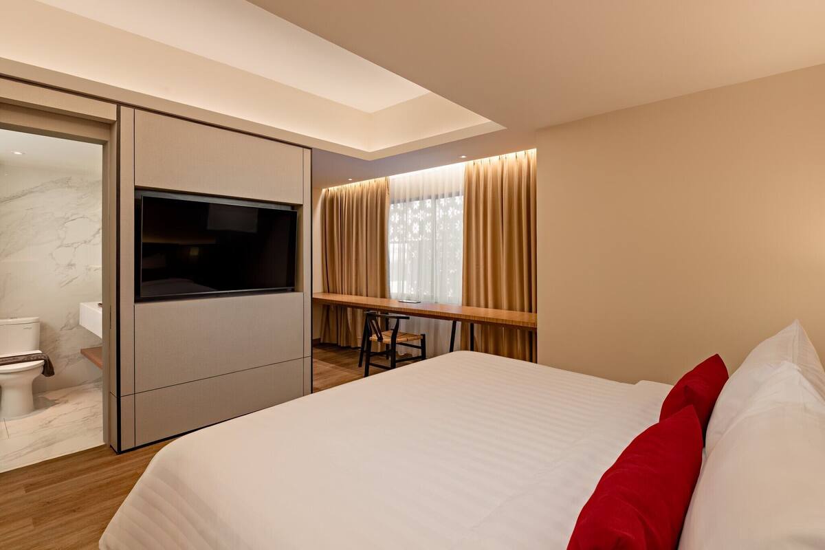 A modern hotel room with a large bed, a desk with two chairs, a television, and a sliding door leading to a bathroom.
