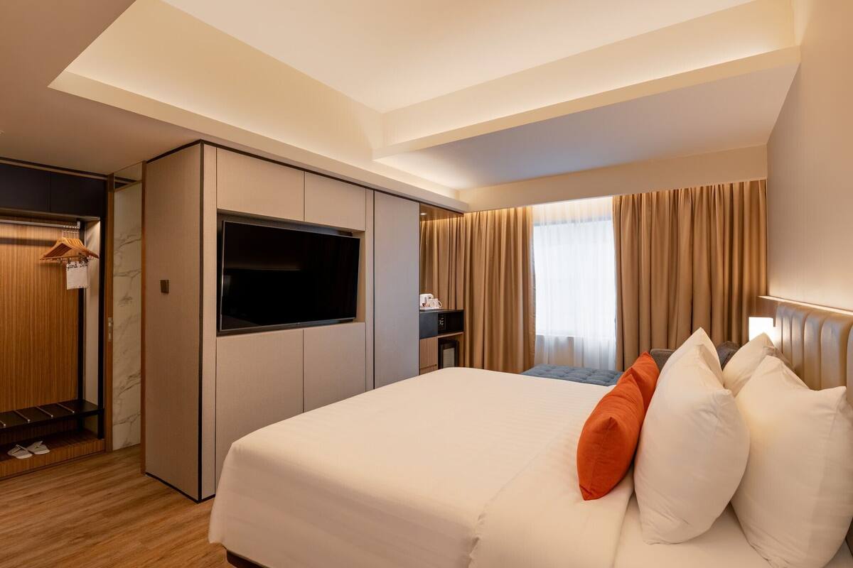 A hotel room with a large bed, a TV, a desk with a chair, a wardrobe, and a window with curtains.