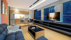 Een moderne woonkamer met een bank, salontafel en televisie. Er is een keukenhoek met een wasmachine en een eettafel.