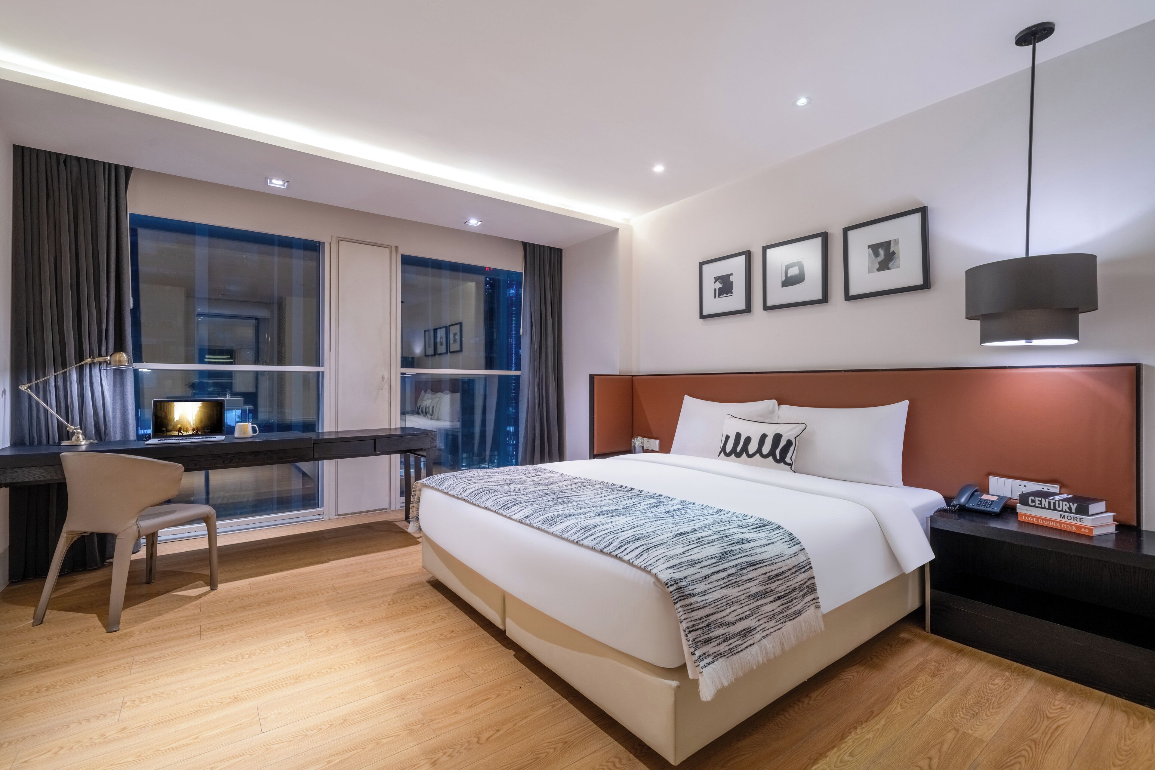 A modern hotel room with a large bed, a desk with a chair, a window with curtains, and a nightstand with a lamp.