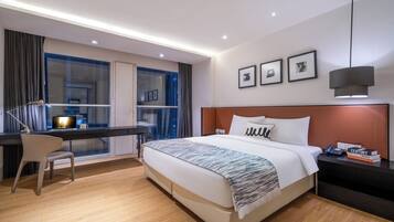 A modern hotel room with a large bed, a desk with a chair, a window with curtains, and a nightstand with a lamp.