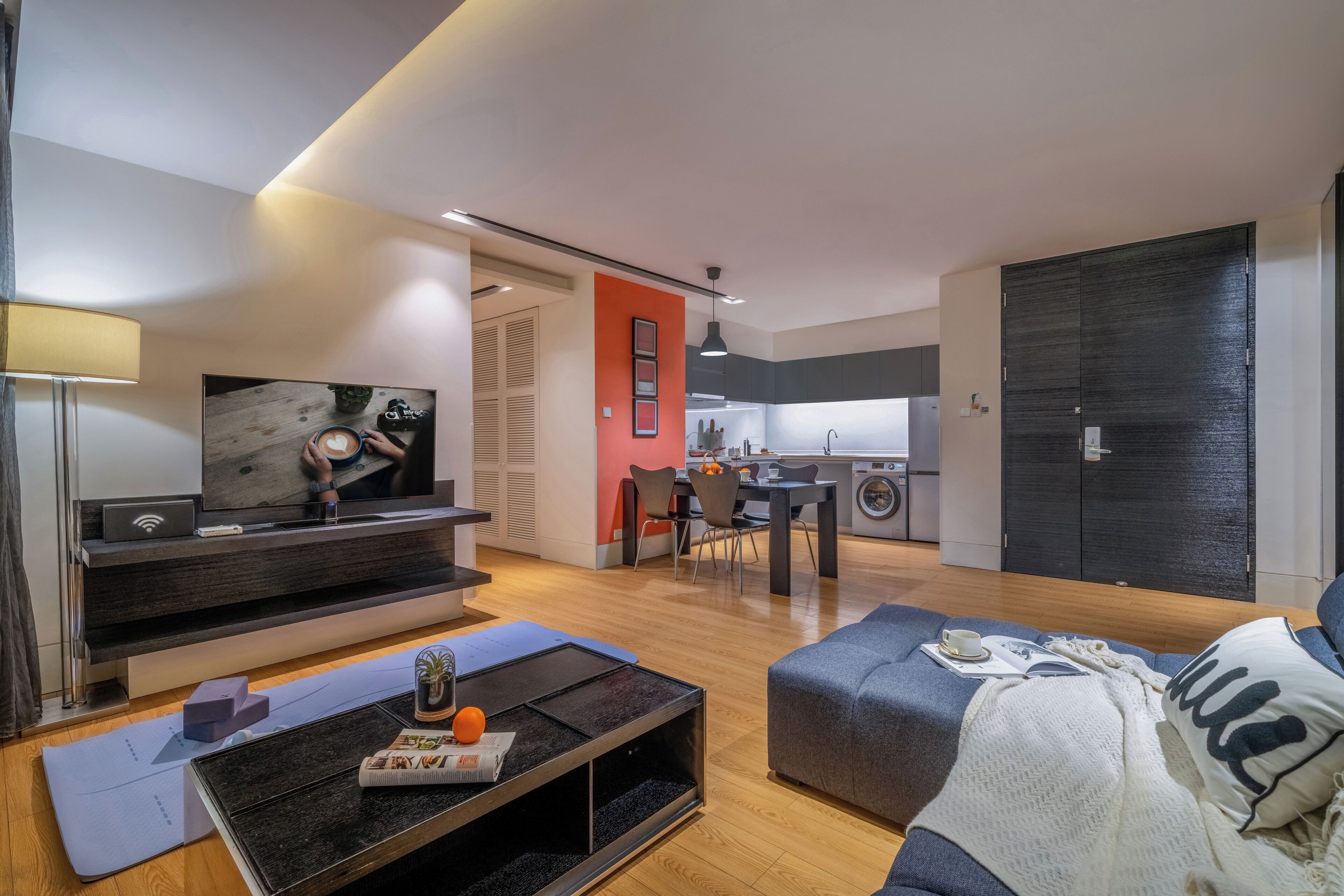 Una sala de estar moderna con un televisor de pantalla plana, una mesa de centro y un sofá. Hay un comedor con una mesa y sillas, y una cocina al fondo.