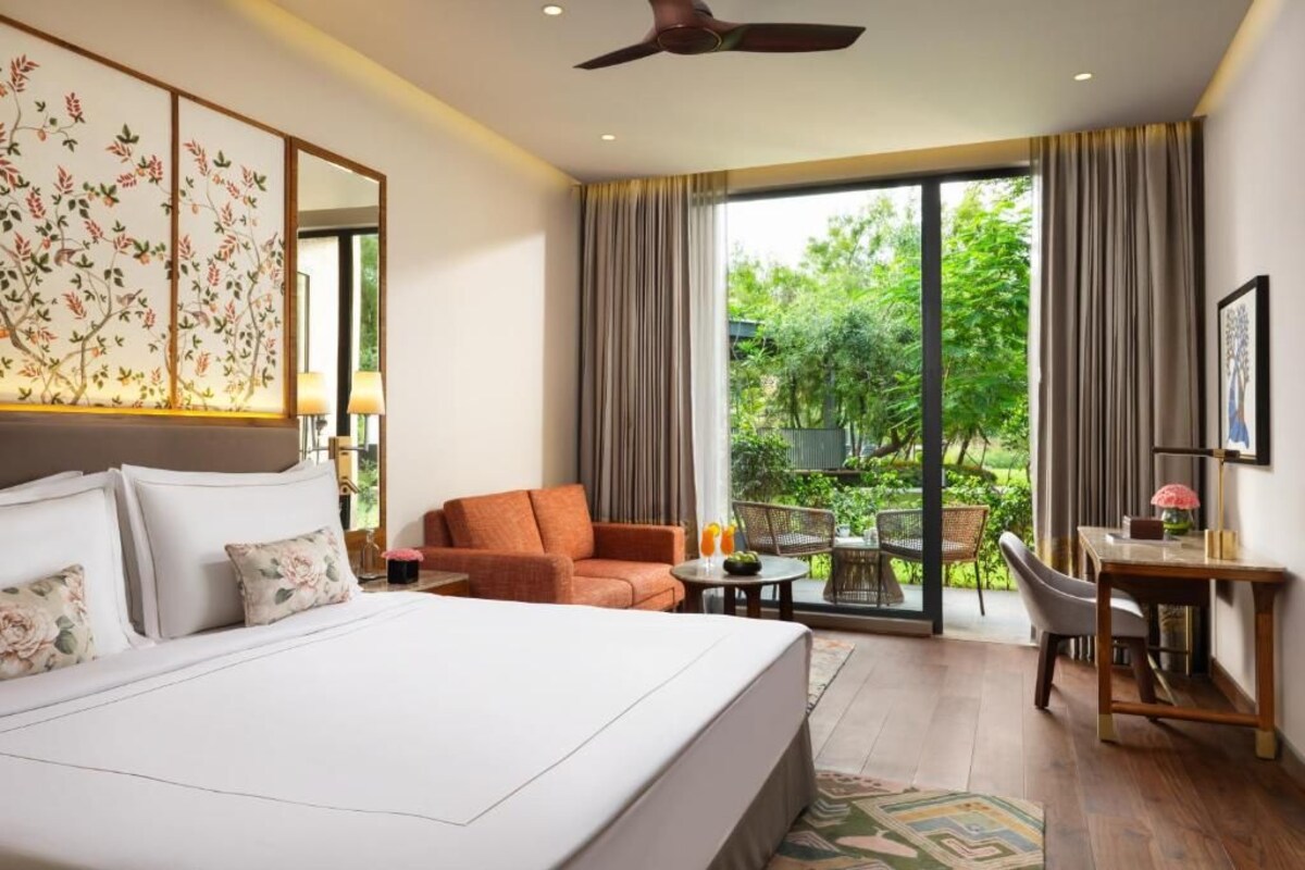 A hotel room with a large bed, a desk, a chair, and a view of greenery through the windows.