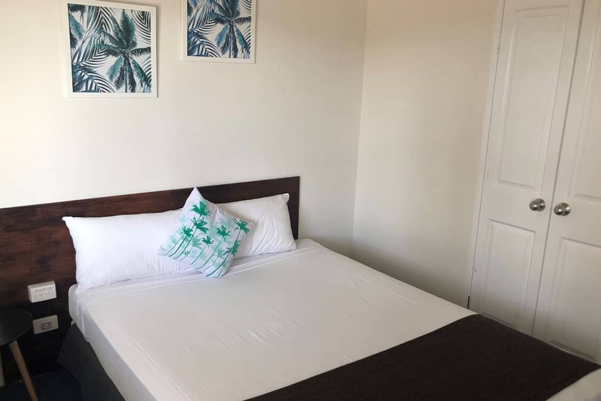 A neatly made bed with white linens and a single decorative pillow, a wooden headboard, and two framed tropical leaf artworks on the wall.