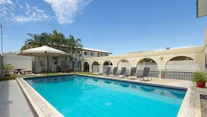 A swimming pool with lounge chairs and a covered area, surrounded by a building with arched windows and a tiled pool edge. - Coral Sands Motel (Mackay)