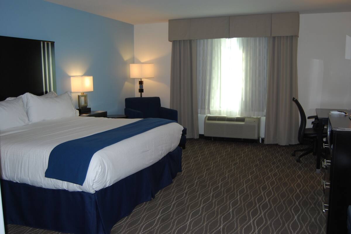 A hotel room with a large bed, two bedside lamps, a desk with a chair, a window with curtains, and a radiator.