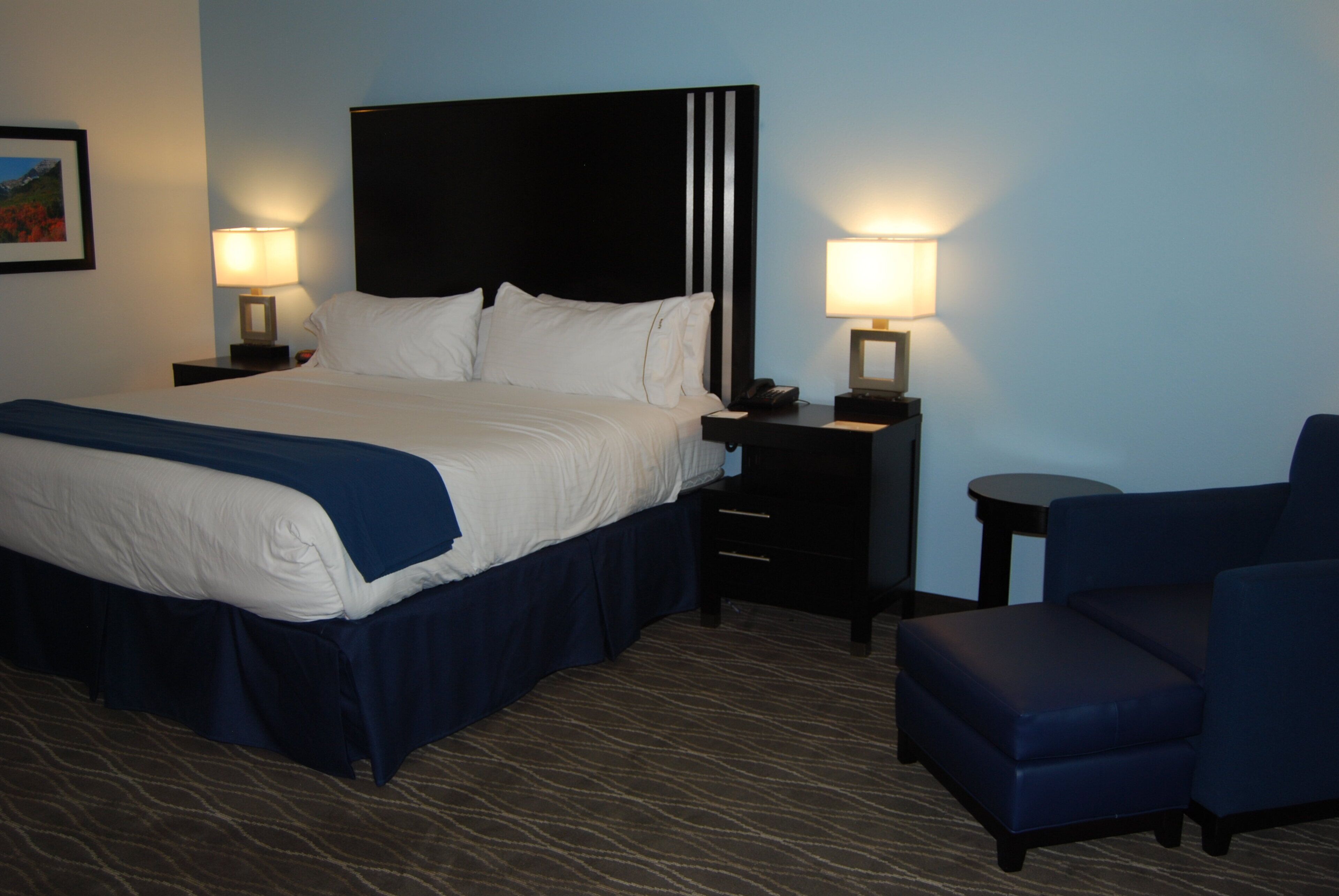 A hotel room with a large bed, two bedside tables with lamps, a chair, and a carpeted floor.