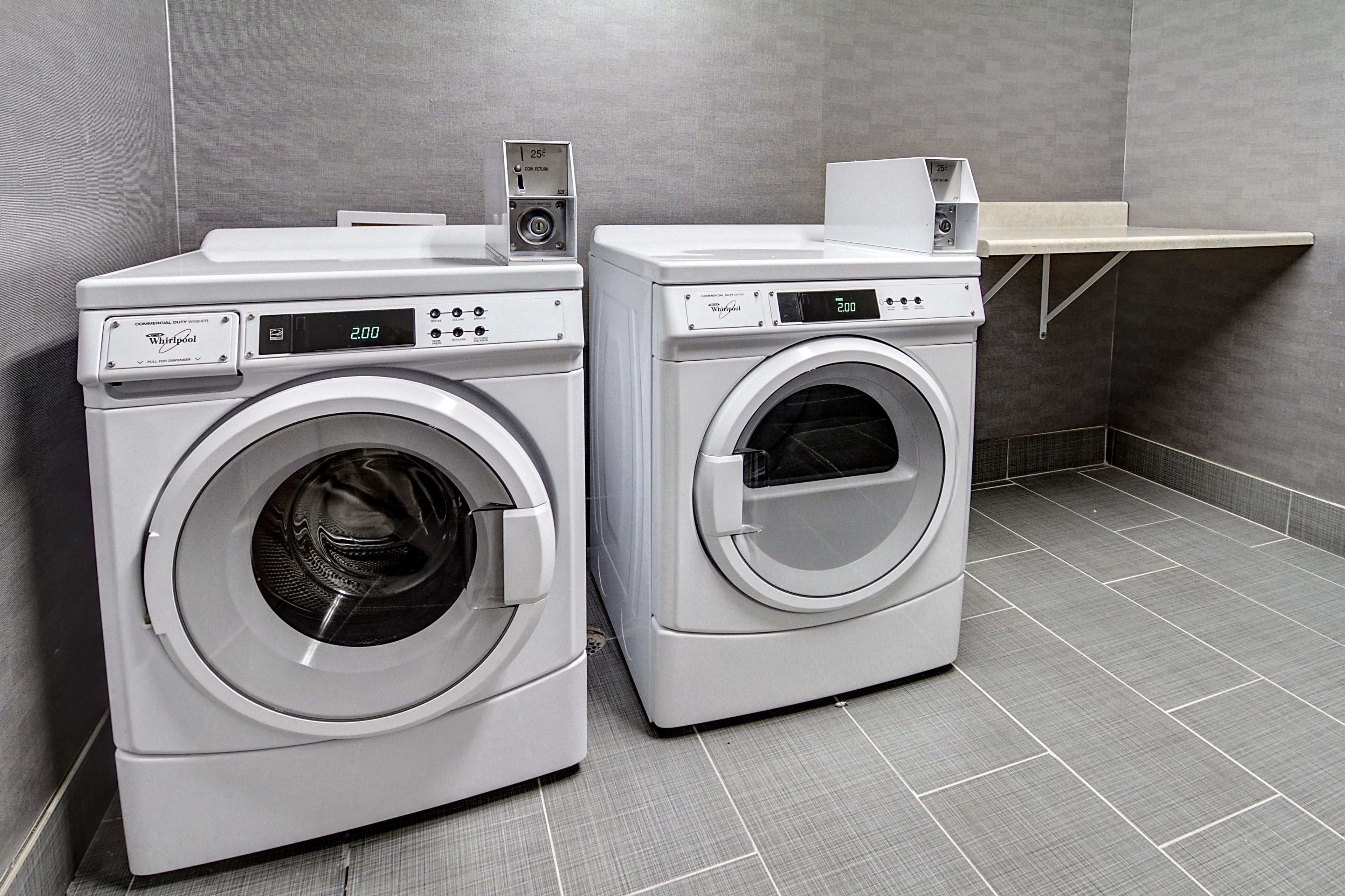 Two commercial duty washers in a utility room with grey tiled flooring and a grey wall.