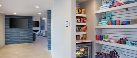 A hotel lobby with a seating area, a wall-mounted TV, and a snack bar with various food items.